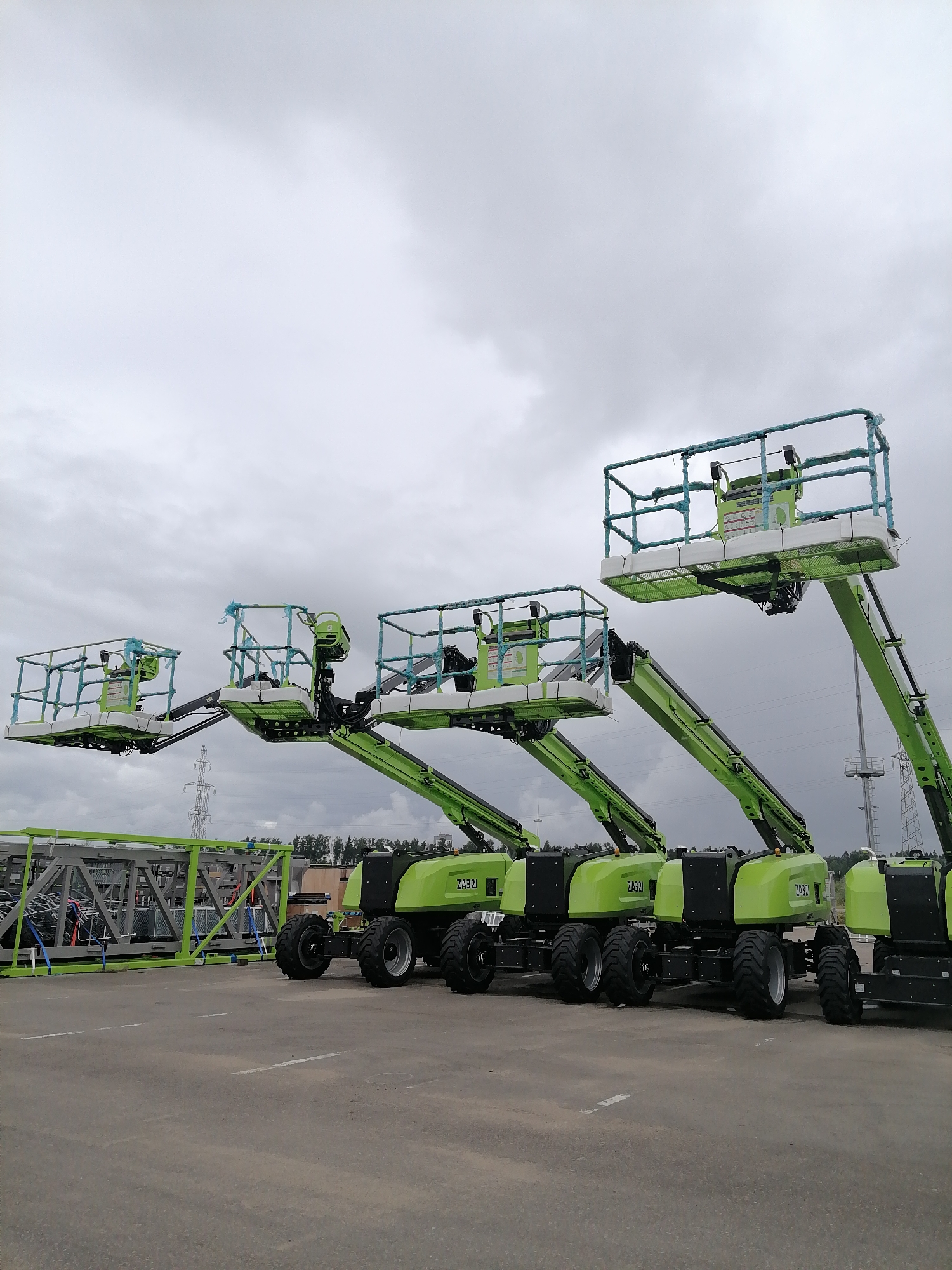The image shows a row of green aerial work platforms parked on a paved lot. The platforms are extended, with their baskets raised and positioned in a diagonal line across the frame. Each platform has a white basket with blue netting around the railing. The bases of the platforms are also green with black accents, and some have visible wheels. In the background, there is a cloudy sky, power lines, and trees. To the left, there is a large metal structure, possibly a scaffolding or equipment rack. The scene appears to be outdoors in an industrial or construction setting. The sky is overcast, suggesting a cloudy or potentially rainy day. The time of day is indeterminate due to the overcast conditions. The ground is paved with white lines marking parking or driving lanes. Visible text on the platforms includes "ZA321J". The overall impression is one of equipment being stored or staged for use.