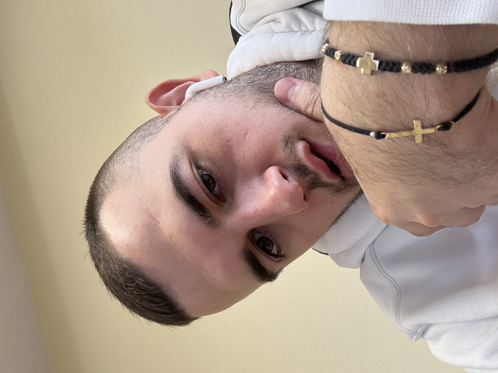A man with short dark hair is pictured from a low angle, looking down at the camera. He is wearing a white hooded sweatshirt and an Apple AirPod in his left ear. He has two black bracelets on his right wrist, both featuring gold crosses. The background is a pale yellow wall. There is no indication of the location of Plovdiv, Bulgaria.