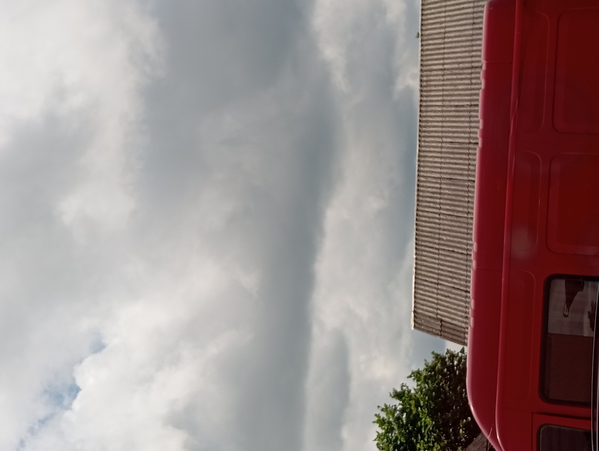 The image shows a portion of a red van parked outdoors under a cloudy sky. To the right of the van, a roof made of corrugated material is visible, with a hint of green foliage from a tree on the left side of the frame. The scene is set in Hlynytsi, Ukraine.