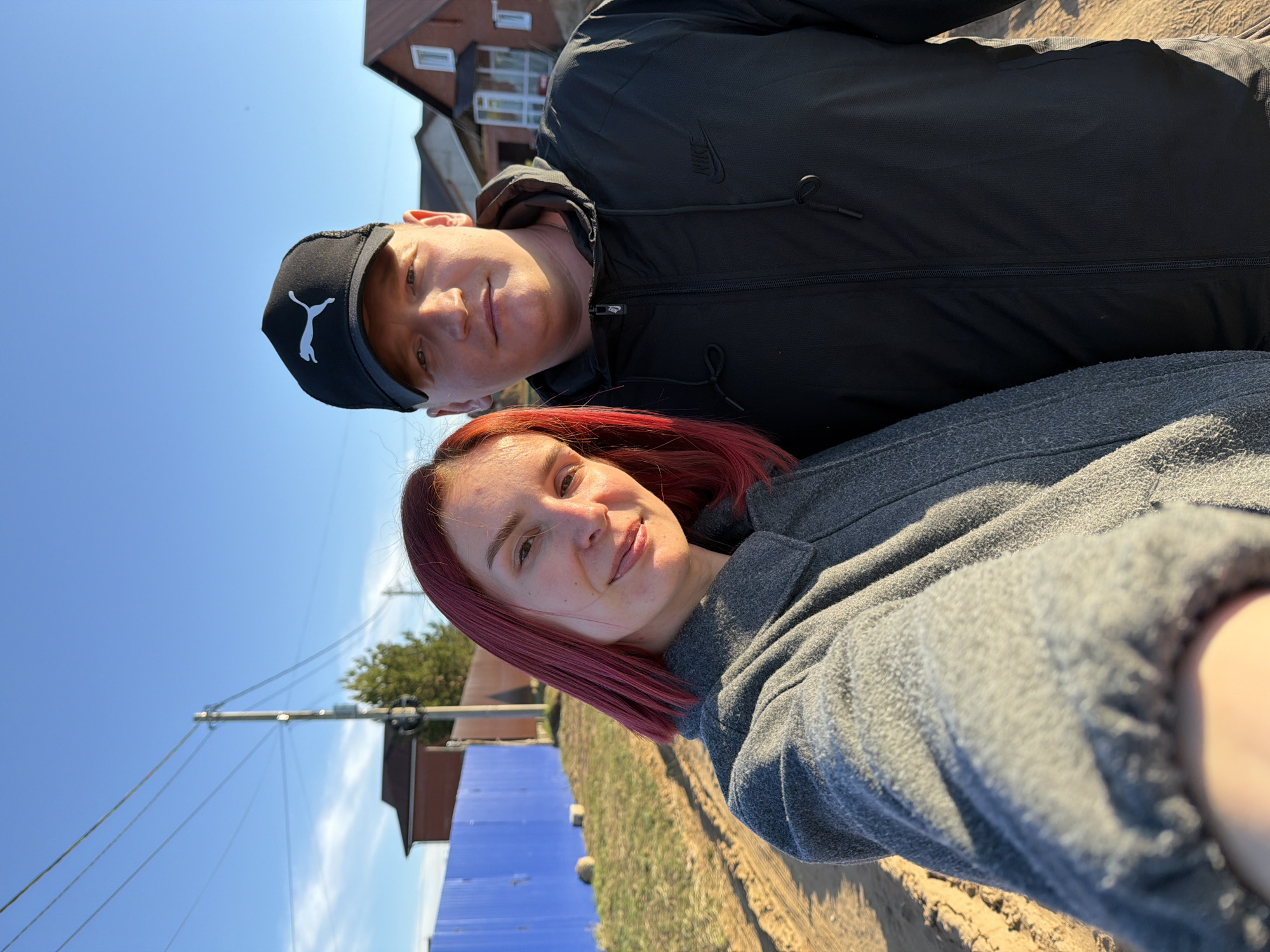 Two people are in the image. The person in the foreground has red hair and is wearing a grey top. The person in the background is wearing a black jacket with a Nike logo and a black Puma cap. The background shows a clear, blue sky, a building, a tree, a blue fence, and telephone poles with wires. The location may be Chaikovsky, Russia.