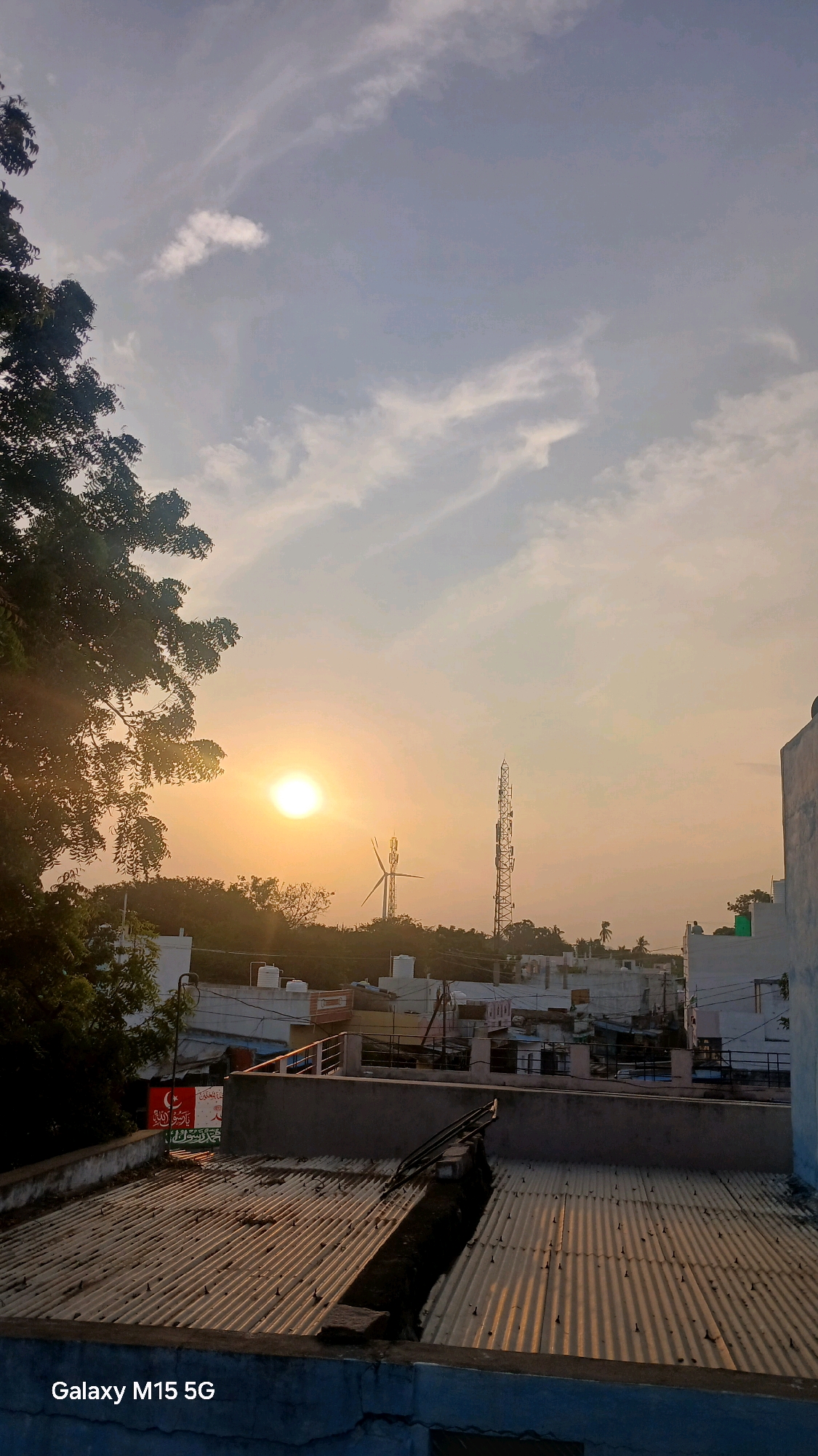 An outdoor, vertical, eye-level, full shot captures a hazy sunset over the city of Vajrakarur, India. The sun is a bright yellow orb in the upper left quadrant, surrounded by a diffuse glow. Wispy white clouds dot the sky. In the midground, silhouetted trees and buildings form a horizon line. A tall cell tower stands prominently to the right of a wind turbine. Rooftops with corrugated metal sheeting are visible in the foreground and midground. A portion of a white building with a blue trim is on the far right. The scene is illuminated by the warm light of the setting sun. The text "Galaxy M15 5G" is visible in the lower left corner. No people are visible in the frame.