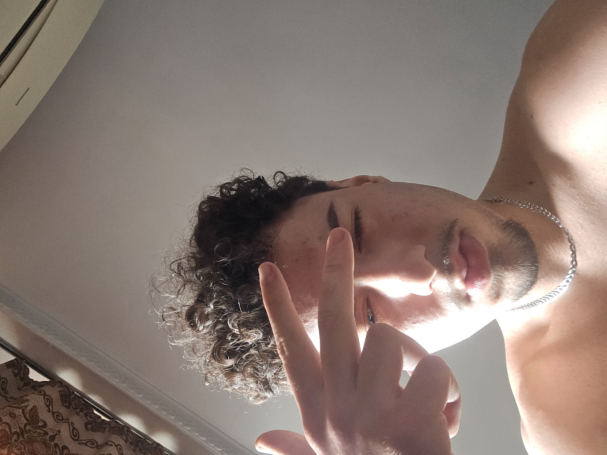 A young male with dark, curly hair, a short beard, and a mustache is depicted in a medium close-up, facing the camera. His right hand is raised, with the index finger extended and thumb up, forming a "finger gun" gesture pointed at his right temple. His eyes appear partially closed or squinting, and his lips are slightly parted. He is shirtless, revealing his bare shoulders and upper chest, and wears a silver chain necklace. Strong directional light illuminates his face and right shoulder, casting shadows. In the upper left background, a portion of a patterned curtain or fabric is visible, along with white ceiling molding. A light-colored wall occupies the majority of the background. The upper right background shows what appears to be the edge of an air conditioning unit or vent. The specific location within Sofia, Bulgaria, cannot be determined from the visual information present in the image.