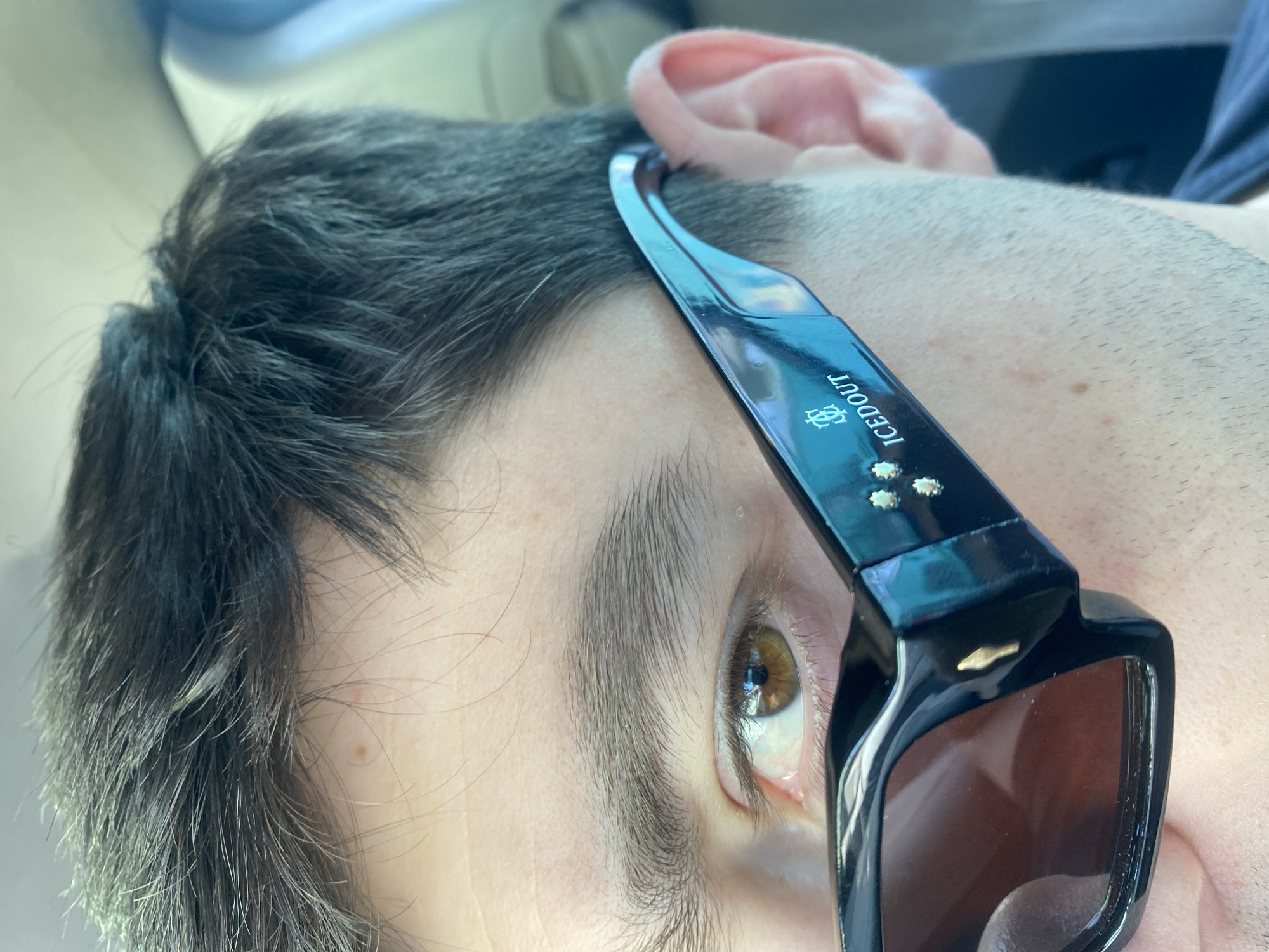 The image shows a close-up view of a person's face wearing sunglasses. The sunglasses are black with the word "ICEDOUT" printed on the temple. The person has dark hair, an eyebrow above their right eye, and hazel eyes. The background is blurry and out of focus. There is no information to discern the location of the image.
