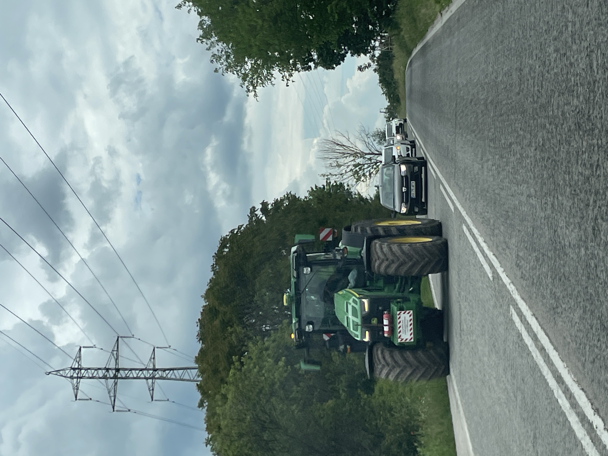 The image shows a green John Deere tractor driving on a narrow asphalt road with double white lines in Kamenar, Bulgaria. There are several cars lined up behind the tractor. Power lines and trees are visible on the left side of the road. The sky is cloudy.