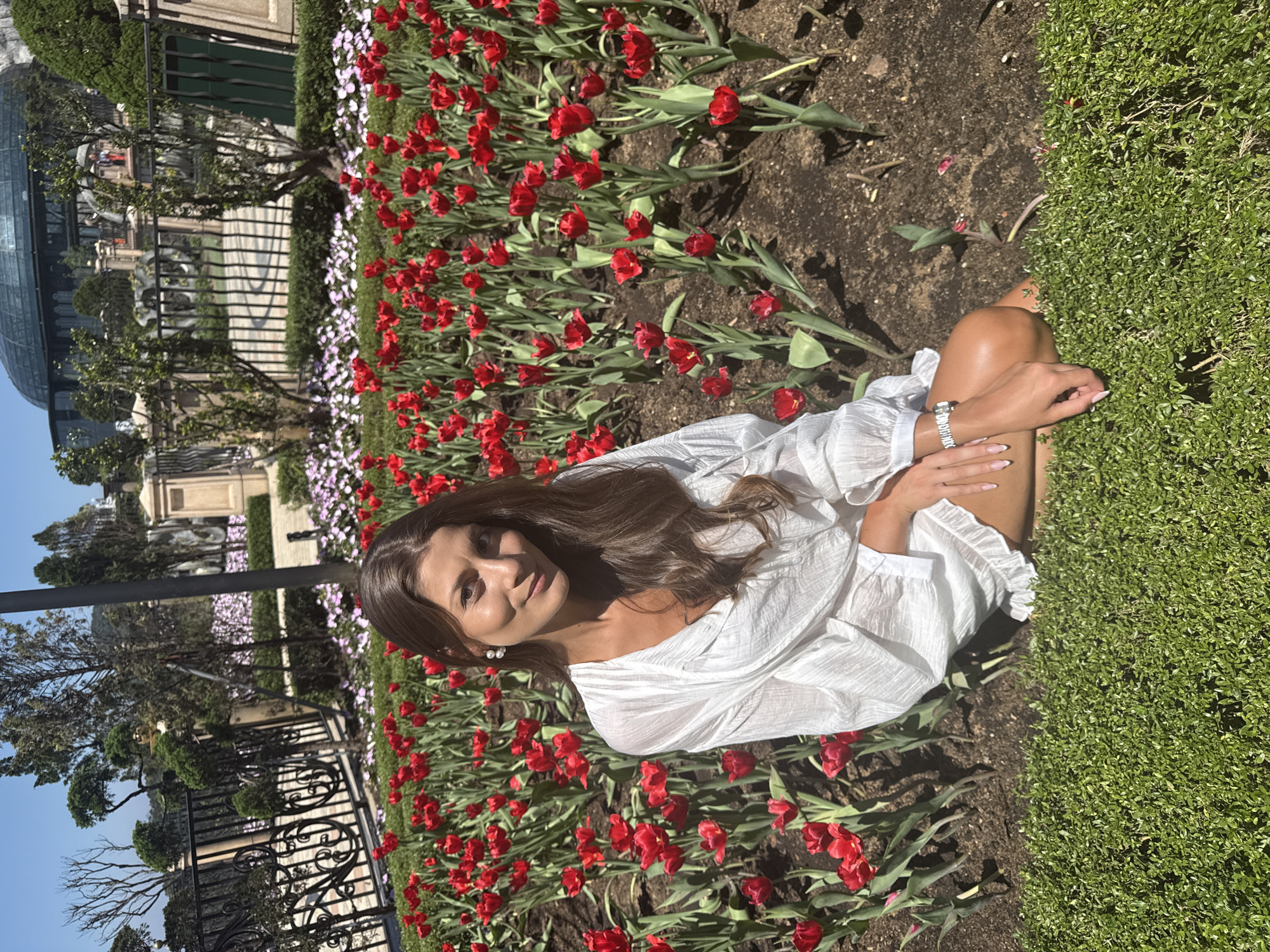 A woman in a white dress sits in a flowerbed of red tulips and grassy groundcover. In the background, there is a fence, a building with a glass dome, and other greenery. The photograph appears to have been taken at Ba Na Hills in Đà Nẵng, Vietnam, based on the architecture and scenery. The woman is looking at the camera. She is wearing a bracelet.