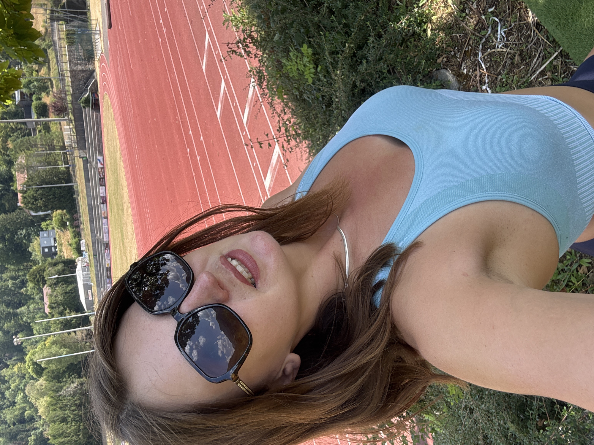 A woman with brown hair and sunglasses is standing on a grassy area next to a running track. The track is red with white lines, and there are stadium seats in the background. Trees and buildings are visible on a hillside behind the track. The woman is wearing a light blue sports bra and has a silver necklace on. The sunglasses reflect the sky and trees. The scene appears to be an outdoor athletic facility. The location is the Stade de la Pégoudière in Saint-Claude, France.