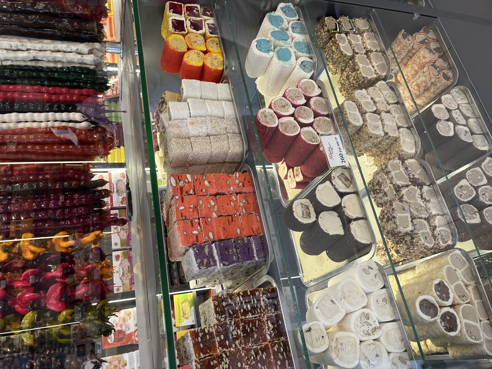 The image displays a vibrant and abundant selection of Turkish delight and other confections arranged in glass display cases. The items are primarily food products, featuring a wide variety of colors, shapes, and textures.
In the left section of the image, several long, rod-shaped confections are stacked vertically. These appear to be made of a chewy, possibly fruit-based material, with some displaying a glossy, deep red color, others a vibrant orange, and some a dark brown. Interspersed among these are lighter colored items, some white and others a deep green.
Moving to the right, the display cases are filled with more traditional Turkish delight squares and rolls. One tray contains square pieces of Turkish delight in bright orange and purple hues, studded with nuts and seeds. Another tray features rolls of Turkish delight coated in what looks like shredded coconut. Further to the right, there are rolls of lighter colored Turkish delight, some coated in chopped nuts and others appearing to be plain. There are also smaller, bite-sized pieces of Turkish delight, some with darker fillings or coatings, and others with a white, powdery exterior.
The confections are meticulously arranged, suggesting a retail setting, likely a confectionery shop or market stall. The lighting appears bright and even, indicating an indoor environment, possibly with artificial lighting. The overall impression is one of sweetness, variety, and visual appeal, inviting customers to browse and purchase.
Visible text includes a price tag in Russian, "260 руб/100 гр", indicating "260 rubles per 100 grams." The location context of Sochi, Russia, further confirms the setting as a place where such delicacies are commonly sold. No people are visible in the image.