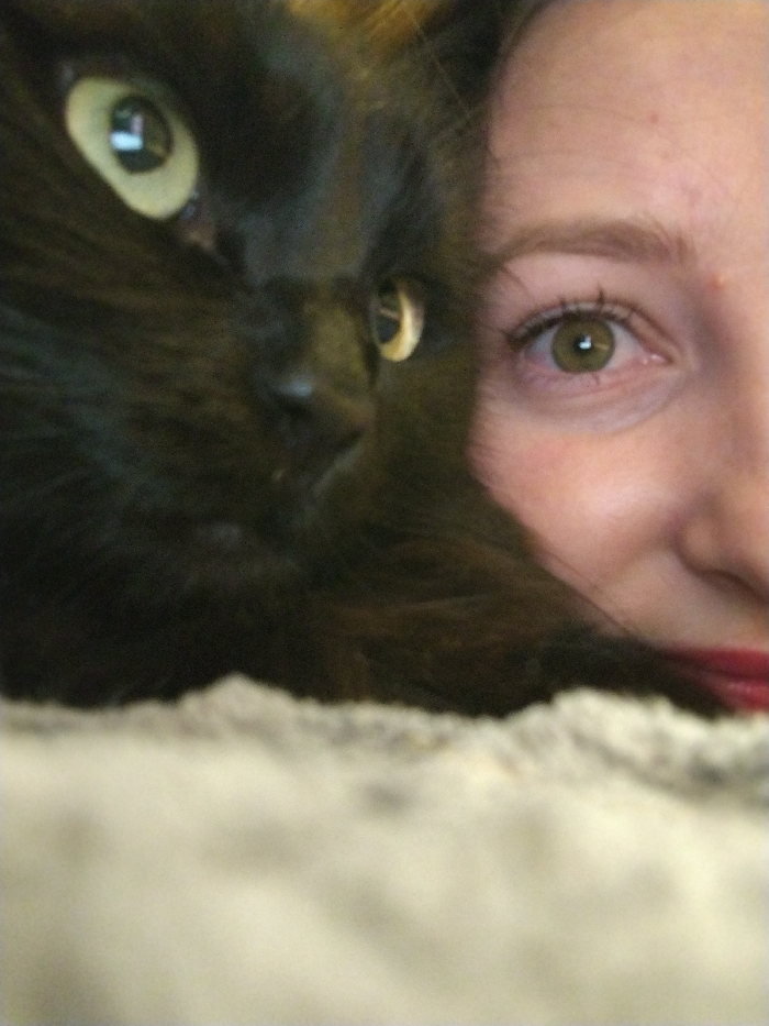 An extreme close-up photograph captures a black cat positioned intimately beside a human female's face, located in Chaikovsky, Russia. The cat occupies the left side of the frame, showcasing its left profile with prominent light green-yellow eyes, each exhibiting distinct reflections of a light source. Its dense black fur covers its head, with its nose and muzzle visible. To the right, a segment of the human female's face is visible, including her right green-hazel eye with visible eyelashes and a light reflection, a natural brown eyebrow, and a portion of her upper cheek and nose. Her lower lip, partially visible, is coated with red lipstick. Brown hair is present, bordering the human's face. The foreground is out of focus, revealing a light-colored, textured material, likely fabric. The background remains indistinct.