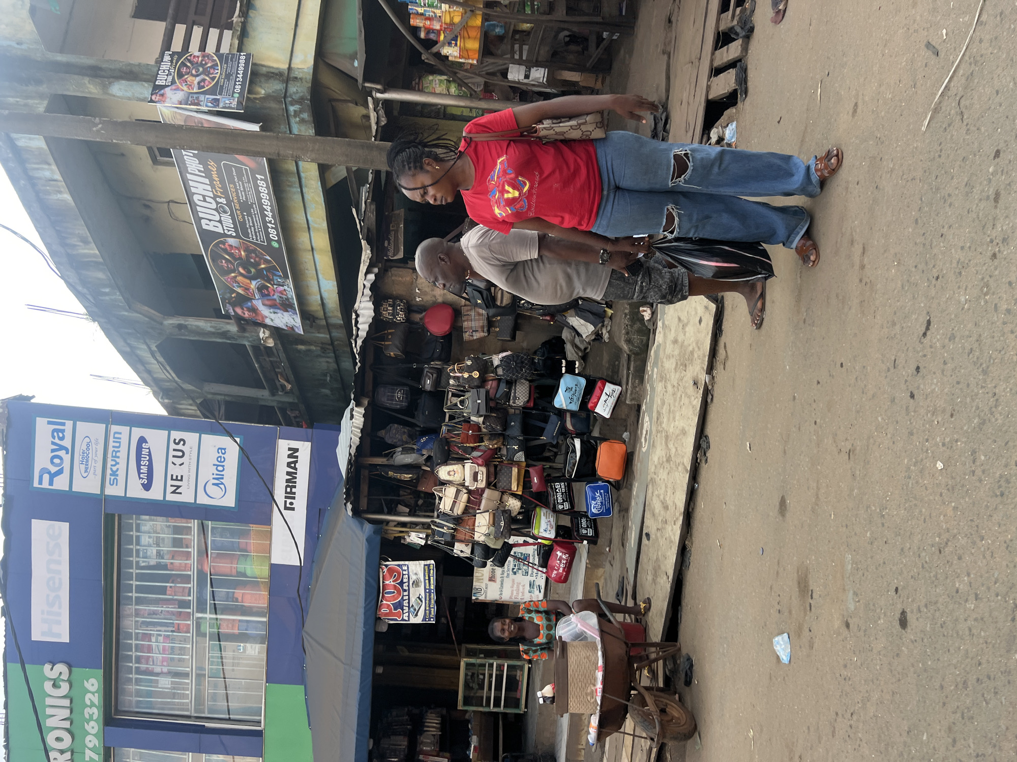 This media file captures a street scene in Nkpor, Nigeria, likely during daylight hours under overcast skies. The foreground is dominated by a busy street with several people. A woman in a red t-shirt and ripped blue jeans walks past a stall selling handbags. Next to her, a man in a grey t-shirt and patterned shorts is also walking. Behind them, a stall is laden with a variety of handbags, displayed on racks and shelves. A woman sits on a cart near the stall, smiling at the camera.
Further in the background, a brightly lit electronics store, "Hisense," is visible with signage that includes brands like "Royal," "Haier Thermocool," "Skyrun," "Samsung," "Nexus," "Midea," and "Firman." Above the street level, the facade of buildings shows various shops and advertisements, including a prominent sign for "BUCHI PHOTO STUDIO & Frames" with a phone number. The overall impression is of a vibrant and bustling market or commercial area with a mix of retail shops and street vendors. The presence of multiple advertisements and the variety of goods suggest a commercial hub.