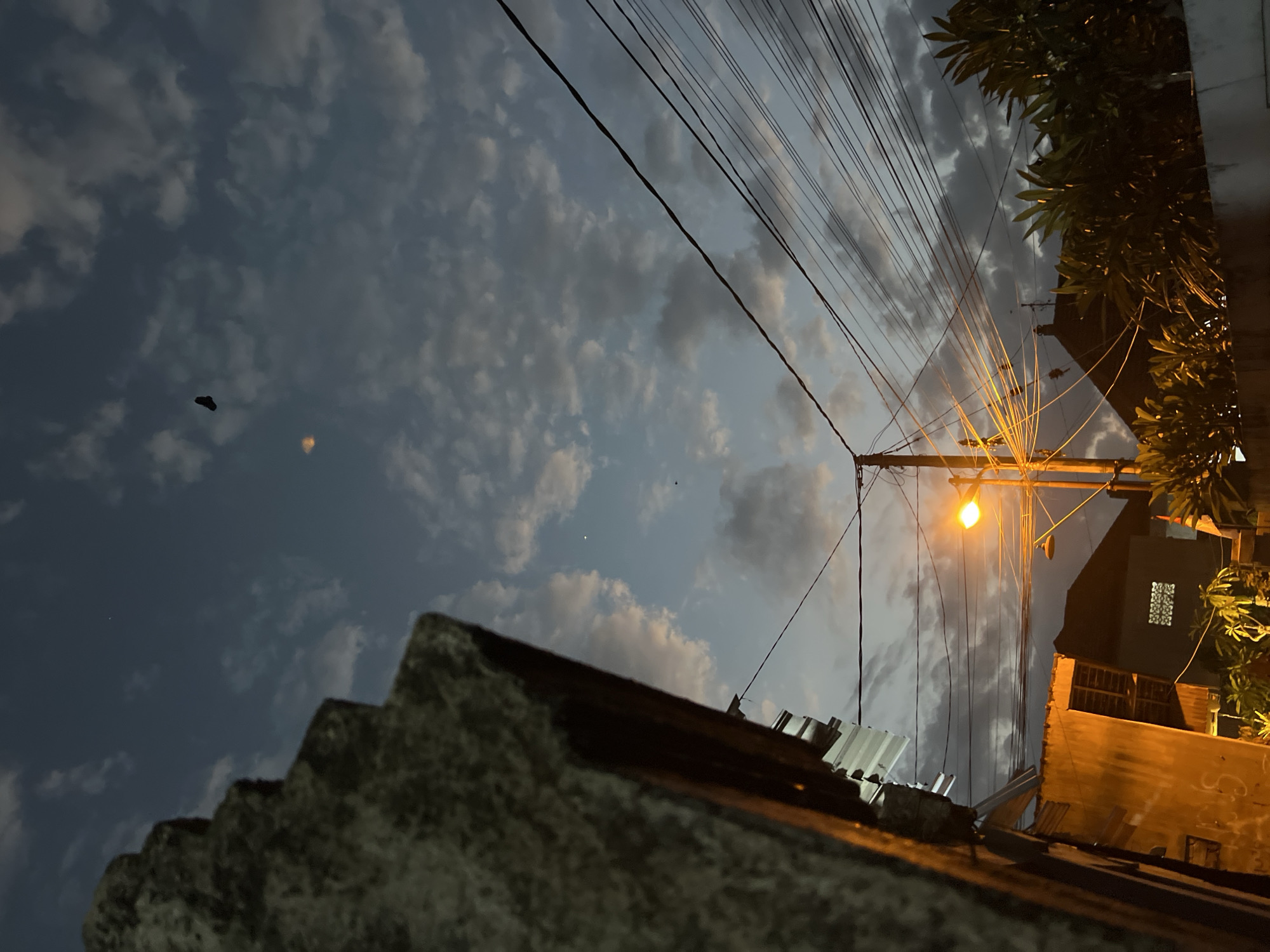 The image captures a twilight scene in Denpasar, Indonesia, likely from a residential area. In the foreground left, a textured stone structure with a corrugated metal roof is visible, illuminated by a warm, orange light. To the right, a street lamp casts a bright yellow glow on tangled power lines that crisscross the frame. Behind the power lines, several houses with dark roofs are visible, one with a small, patterned window. Lush green foliage in the foreground right partially obscures a concrete wall. The sky is a deep blue with scattered, wispy clouds tinged with orange and grey from the setting sun. In the upper portion of the sky, a dark, kite-like object is suspended, and a small, faint light, possibly a planet or distant light, is also visible. The overall impression is one of a quiet evening in an urban Indonesian setting, with a mix of natural and man-made elements.