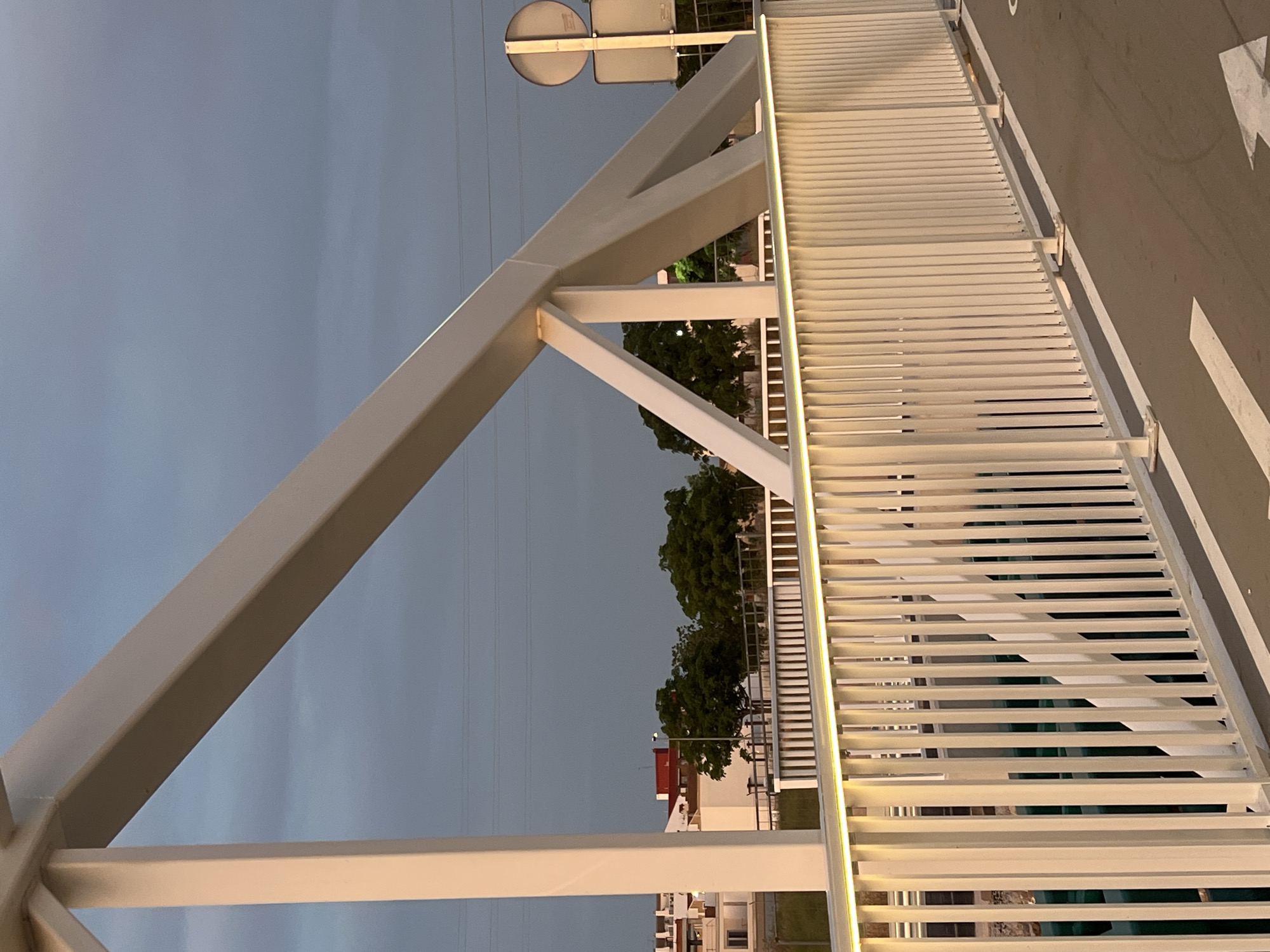 This image depicts a modern, white-painted metal truss bridge located in Alcàsser, Spain, during what appears to be twilight or dusk. The bridge's superstructure features prominent, rectangular cross-section beams arranged in a truss configuration, with an inclined main beam supported by vertical and diagonal struts. A white metal railing runs along the visible side of the bridge, composed of numerous vertical balusters. A warm, yellow LED strip light is integrated along the top edge of this railing, illuminating the structure. Below the railing, a paved path or road surface is visible, marked with white directional arrows, one partially visible pointing towards the lower right.
Through the gaps in the bridge's structure and railing, a body of water, possibly a canal or river, can be seen with a dark greenish hue. Beyond the bridge on the left, white residential buildings with terracotta-colored roofs are visible against the horizon. Clumps of green trees are interspersed between the buildings and the waterway. In the upper background, thin horizontal lines, indicative of overhead power lines, traverse the scene. On the right side of the bridge, the back of a circular road sign and a rectangular sign, both mounted on a metal pole, are partially visible. The sky overhead is a uniform, muted blue-gray, suggesting overcast conditions or the time of day. No people or animals are present, and no specific actions or interactions are depicted.