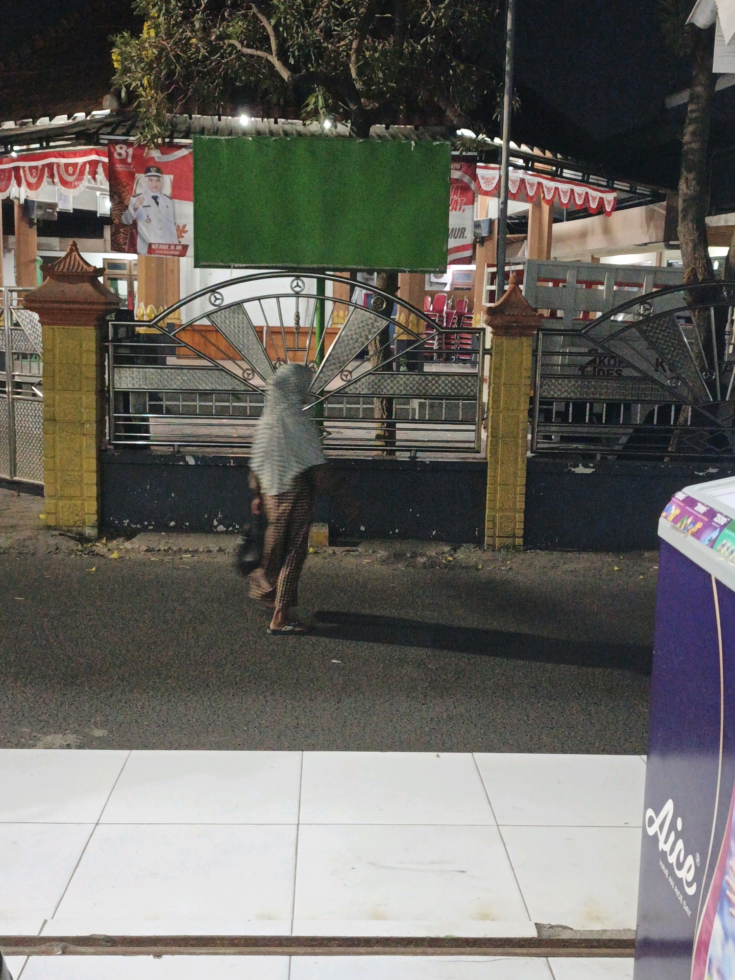 In the city of Ketegan, Indonesia, a person wearing a hijab and patterned pants is walking on the asphalt road at night. They are holding a dark bag in their left hand. To the left of the person, there is a yellow pillar and a decorative metal fence. Behind the fence, a building with red and white awnings is visible. A large green rectangular banner hangs above the fence. To the right of the person, a purple-colored kiosk is partially visible. The ground in the foreground is paved with white tiles. The scene is lit by artificial lights, casting shadows on the road.