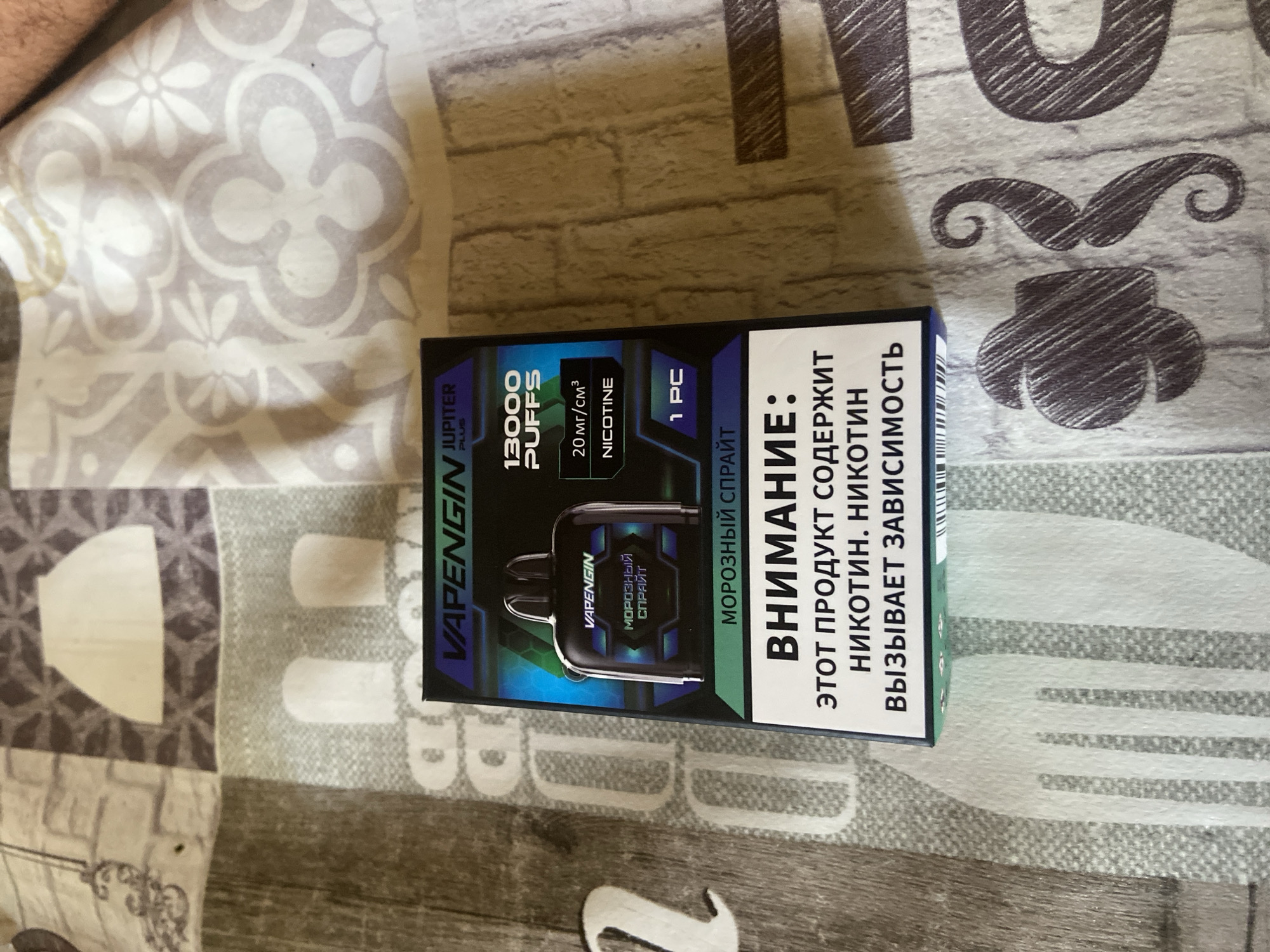 The image shows a close-up of a vape product packaging, likely a disposable vape, lying on a patterned fabric surface. The packaging is primarily dark with blue and green accents. Prominently displayed are the brand name "VAPENGIN" and the product model "JUPITER PLUS". Key information includes "13000 PUFFS", "20 МГ/СМ³ NICOTINE", and "1 PC". There is also text in Russian that translates to "FROSTY SPRITE" and a warning label stating "ATTENTION: THIS PRODUCT CONTAINS NICOTINE. NICOTINE CAUSES ADDICTION." The fabric it rests on has a repeating pattern of floral and abstract shapes in shades of grey, beige, and white. The lighting suggests an indoor environment. There are no visible people or specific landmarks that would place this image in Taganaogsky, Russia.
