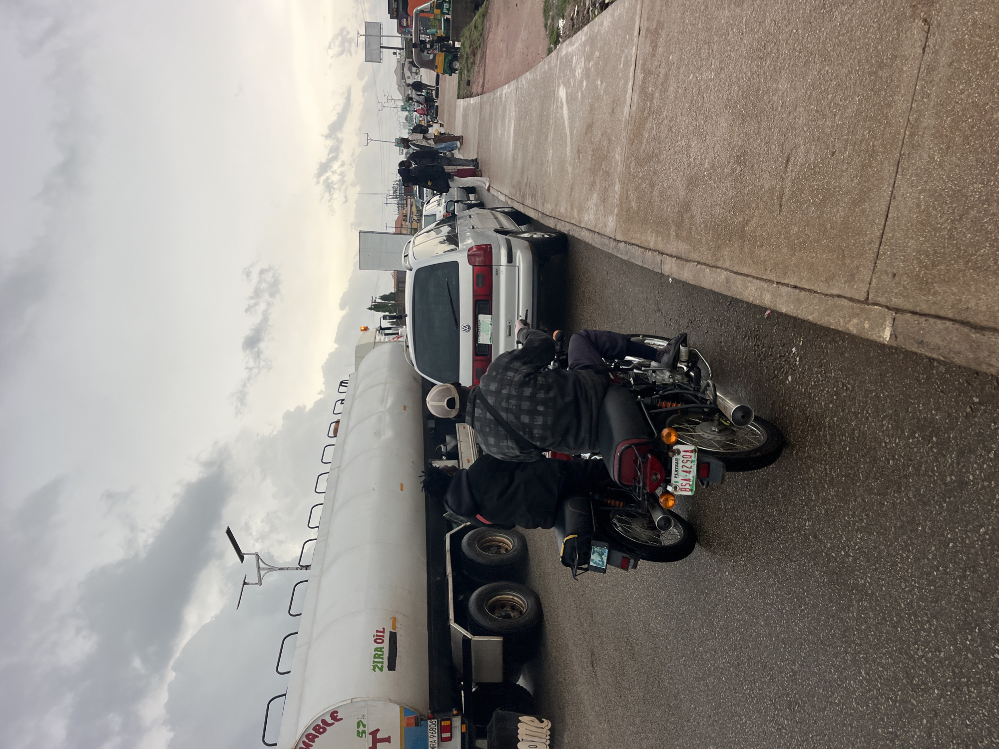 The image depicts a traffic scene with several vehicles and people. In the foreground, two individuals are seated on a motorcycle, facing away from the camera. The rider wears a checkered shirt and a white helmet, while the passenger is dressed in black. Behind them, a white tanker truck, likely carrying oil as indicated by the text "ZIRA OIL" visible on its side, is stopped. Further down the road, a line of cars and other vehicles are also stationary, suggesting a traffic jam. People can be seen standing and walking along the side of the road, which has a concrete sidewalk. The sky is overcast with clouds, indicating it might be a cloudy or rainy day. The overall scene appears to be an urban or semi-urban environment. The license plate on the motorcycle reads "PLATEAU BSA 4250A".