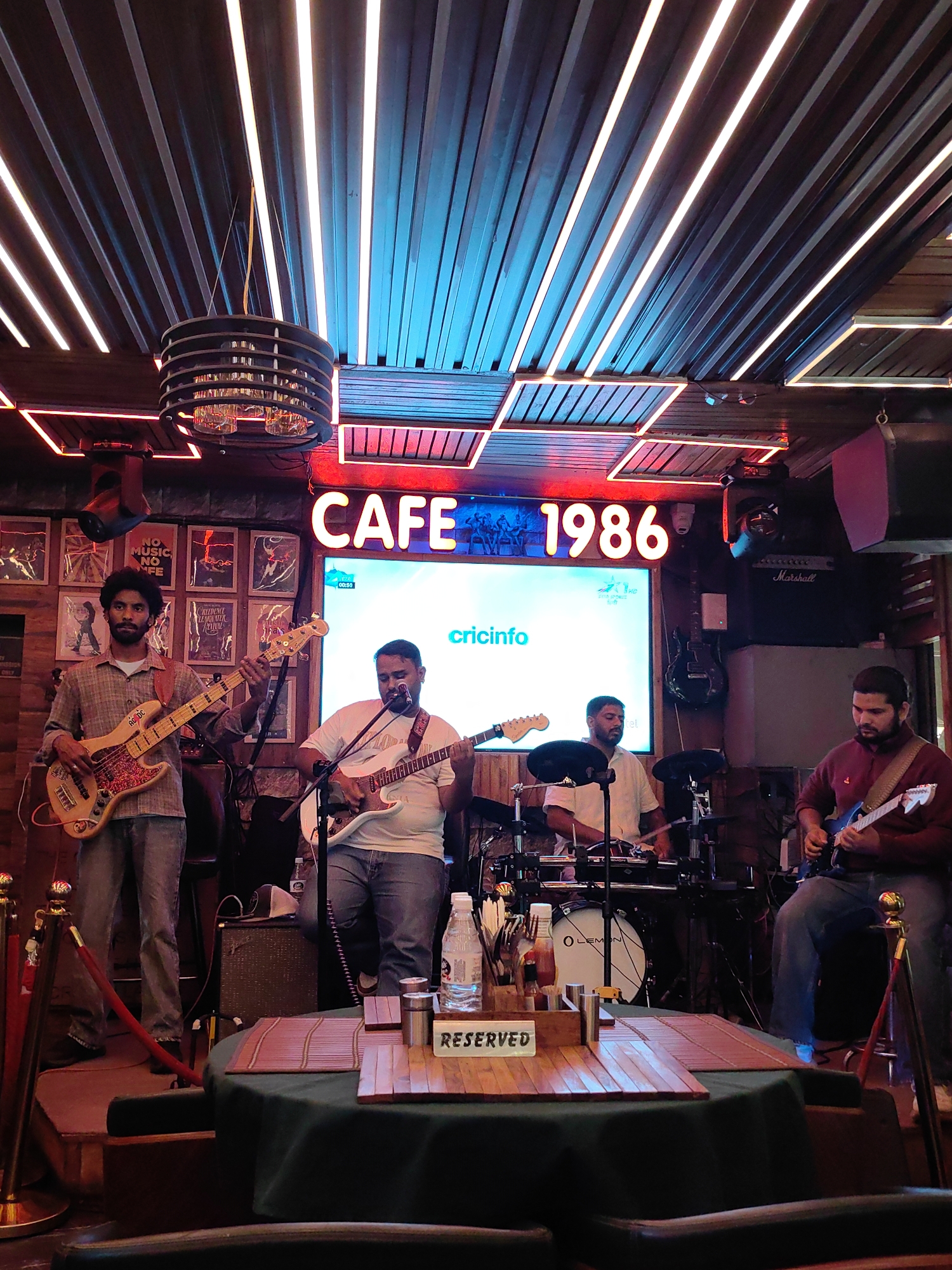 A band of four men is performing on a stage in what appears to be a bar or restaurant. The band members are playing guitars and drums. The stage is decorated with posters and a large illuminated sign that reads "CAFE 1986". The ceiling is adorned with modern lighting fixtures. A table with a "RESERVED" sign is visible in the foreground. The image is taken from a slightly low angle, looking up at the stage.