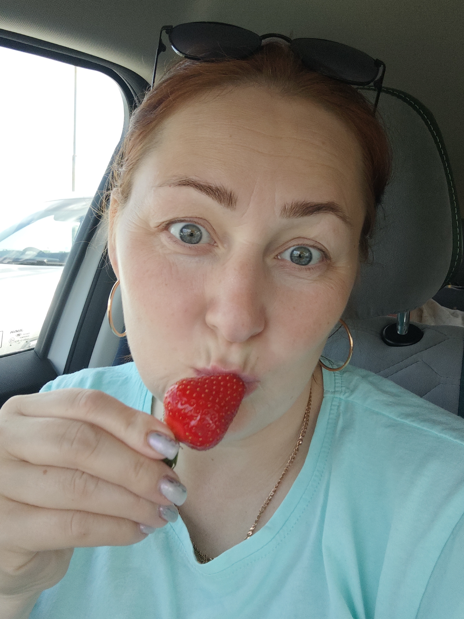 A woman with fair skin and reddish hair, wearing a light blue t-shirt and gold hoop earrings, is holding a ripe red strawberry to her lips in what appears to be a selfie taken inside a car. She is looking directly at the camera with wide eyes. Sunglasses are perched atop her head. A gold necklace is visible around her neck. Her fingernails are painted in a silver, marbled pattern. The car's interior is visible, with a gray patterned headrest and a white car visible through the window. The image was taken in the city of Chaikovsky, Russia.