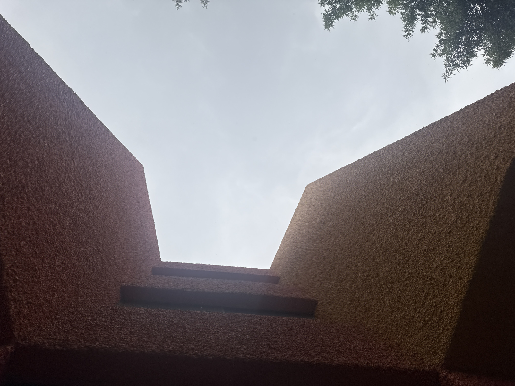The image shows a low-angle view of two brown textured walls converging towards a bright, overcast sky. Tree branches are visible in the upper corners of the frame. The walls appear to be part of a building, possibly a courtyard or entryway. There are two horizontal protrusions on the lower part of the left wall. The location is inferred to be San Andrés Cholula, Mexico.