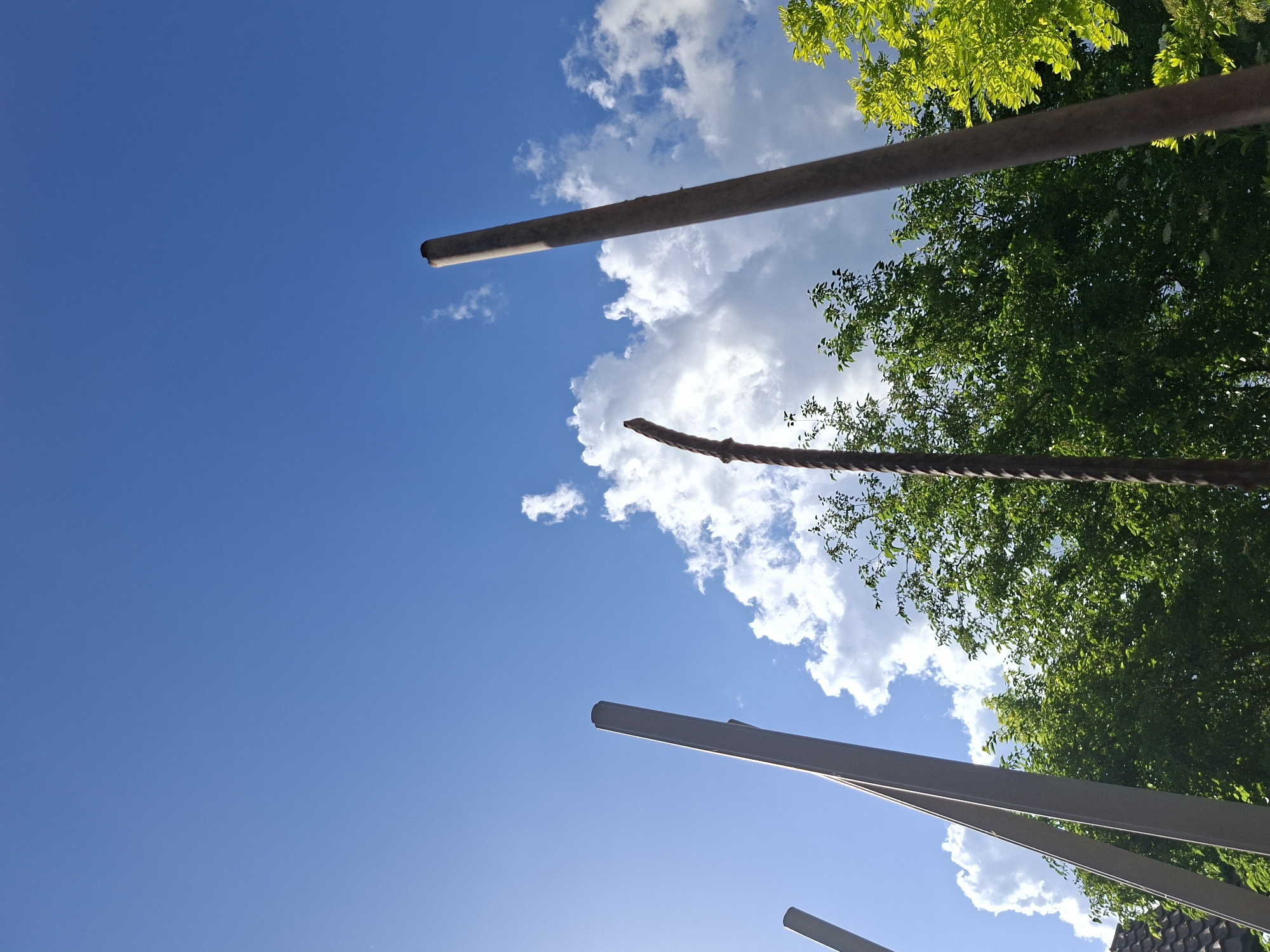 The image depicts a low-angle view towards a blue sky with cumulus clouds, observed between several partially visible structures and foliage. There are tree branches with green leaves present on the right, partially obscuring the sky. Some of the structures appear to be metal poles or supports, with some straight and others curved. One appears to be a thick rope. The scene appears to be outdoors, and the photograph was taken during the day with sunlight. No people are visible. The location, Bojuritsa, Bulgaria, is an additional piece of information.