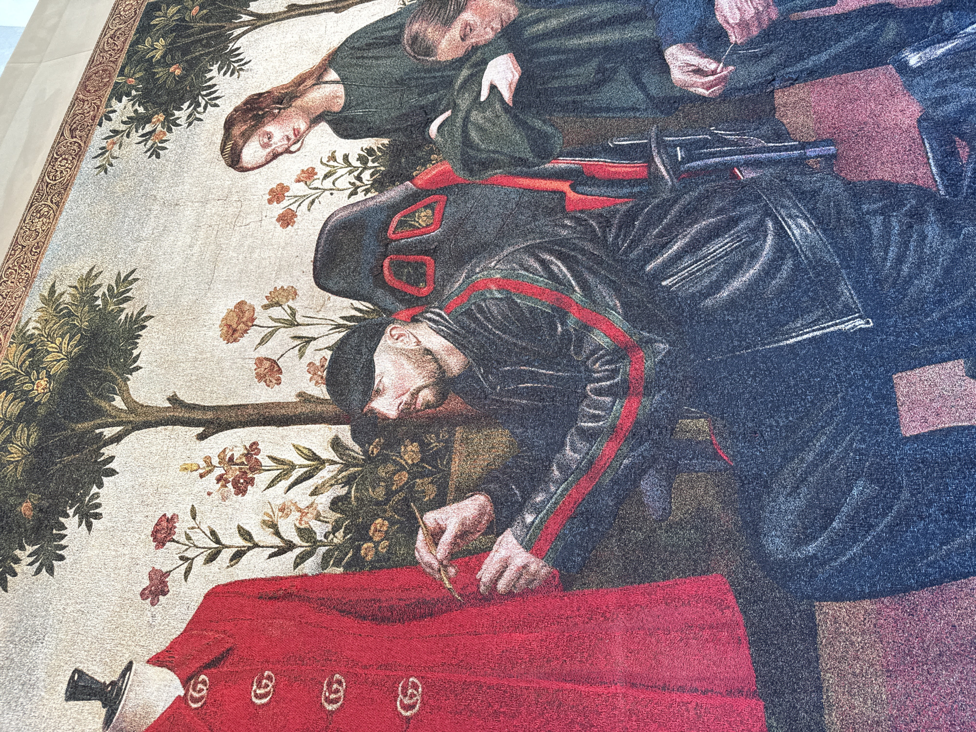 The image depicts a tapestry or wall hanging featuring an artistic interpretation of a scene. In the foreground, a man in a black jacket with red stripes on the sleeves is bent over a red cloth, possibly writing or drawing on it with a small tool. A sword or dagger is partially visible on the left side of the red cloth. Behind him, two figures are lying down, one of whom is a woman with long hair. Behind them there is a tree or foliage with flowers. The tapestry has a decorative border.
While the style is reminiscent of historical art, the man's clothing with the stripes gives it a contemporary, possibly postmodern, twist.
There is no information in the image or context that indicates it is located in Milan, Italy. There are no landmarks or indicators of location. The image cannot be associated with any known location in Milan.