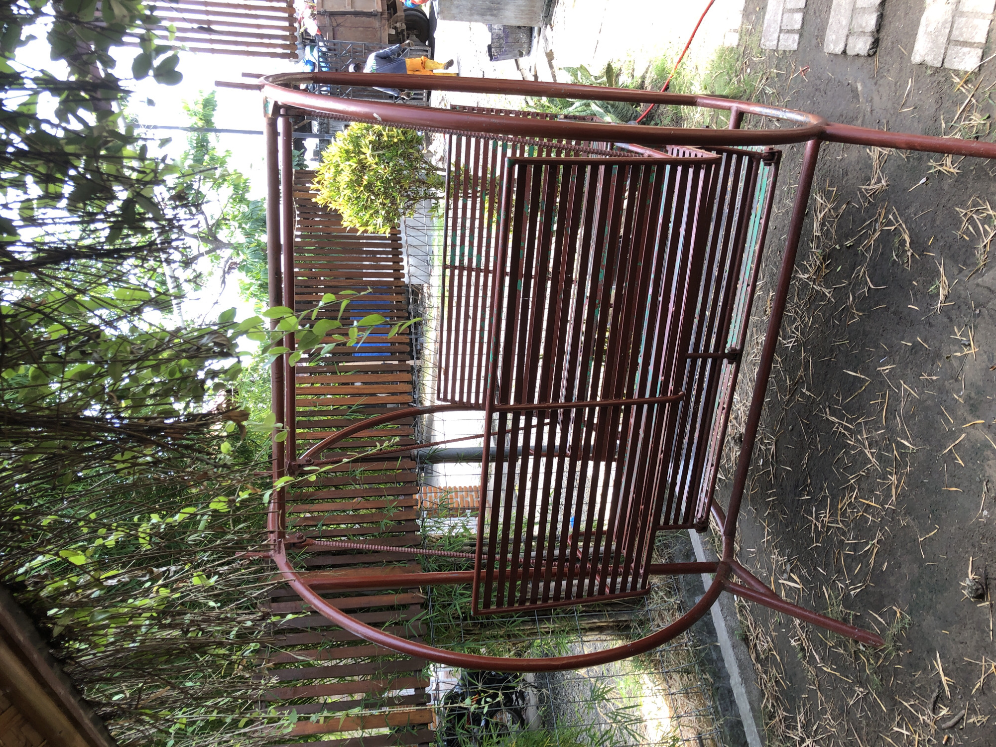 This is a medium-shot, outdoor photograph of a rusty, dark red metal swing set. The swing set has a curved frame with a slatted seat and backrest. It is positioned on a patch of dark soil scattered with dry leaves and grass. Behind the swing set is a wooden fence made of vertical planks, with some green foliage growing in front of it. To the right of the swing set, a small, bushy green plant is visible. In the background, there is a truck and a person wearing an orange shirt and yellow boots. The scene appears to be a backyard or an outdoor seating area in a tropical climate, possibly in Tambonbaroh, Indonesia, judging by the vegetation and the presence of outdoor elements. The lighting suggests it is daytime, with bright sunlight filtering through trees overhead.
