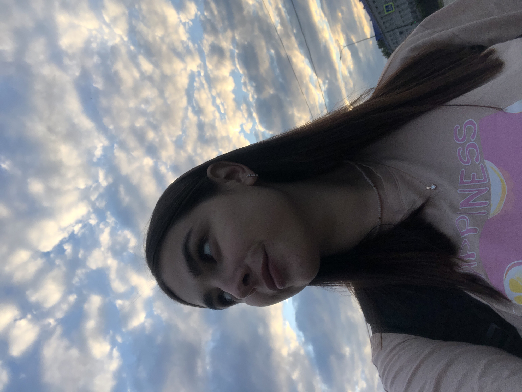 A young woman with dark brown hair is captured in a close-up, low-angle outdoor shot against a backdrop of a dramatic cloudy sky. She is looking upwards and to her left, with a slight smile on her face. Her visible clothing includes a light-colored t-shirt with pink lettering that says "HAPPINESS" and a necklace with a small pendant. She is also wearing an earring in her visible ear. In the background, a streetlamp and buildings can be observed, suggesting an urban setting. The lighting indicates it may be dusk or dawn. The image was taken in Chaikovsky, Russia.