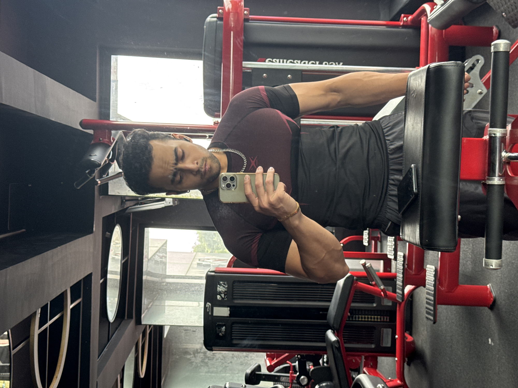 A man is taking a mirror selfie in a gym. He is wearing a burgundy and black t-shirt, gray shorts, and a silver chain necklace. He has a gold bracelet on his left wrist and is wearing black earbuds. He is holding a silver iPhone in his right hand. The gym has red and black equipment. The image is taken in a gym in Kolkata, India.