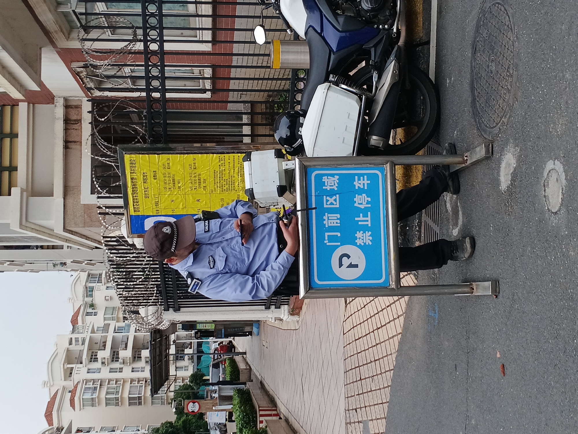 This media file depicts an outdoor scene in Shunde District, China, during daylight hours. The central subject is a person, likely a security guard or police officer, dressed in a light blue uniform shirt with "POLICE" patches on the sleeves and a dark baseball cap showing "C05364". The person is standing next to a blue rectangular sign that features a white 'P' symbol with a diagonal line through it, clearly indicating "No Parking." The sign's text reads "门前区域 禁止停车" (Ménqián qūyù Jìnzhǐ Tíngchē), meaning "Area in front of the gate/door, no parking allowed."
Adjacent to the officer and the sign is a blue and white motorcycle, equipped with white side storage compartments and a helmet resting on its seat. Behind them, a dark metal fence, topped with rolls of barbed wire, separates the street from a building with brickwork and white-framed windows. Attached to this fence is a large yellow sign titled "碧桂花城停车场收费标价牌(陈村管理处)" (Country Garden City Parking Lot Price List (Chencun Management Office)). This sign details the "定价方式:市场调节" (Pricing method: market adjustment) and "收费标准" (Charging standards), listing various parking rates for different times of the day and vehicle sizes.
In the background, several multi-story residential buildings with light-colored facades and reddish-brown tiled roofs are visible. The ground consists of both asphalt roadway, where a manhole cover is visible, and a patterned tiled sidewalk. The scene suggests a structured environment, possibly a residential complex or business entrance, where parking rules and services are clearly communicated and enforced.