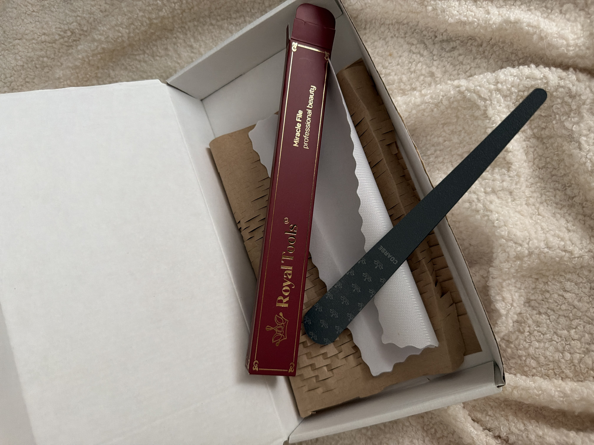 An opened white cardboard box is placed on a fluffy, cream-colored blanket, suggesting a cozy, indoor setting. Inside the box, there are several items related to nail care. A red and gold-embossed box, labeled "Royal Tools" and "Miracle File professional beauty," is positioned prominently. Next to it, a black nail file with a decorative pattern and the word "COARSE" visible on its surface lies on white tissue paper. Crinkled brown paper is used as padding within the box. The image appears to be taken in natural light, possibly during the daytime. There are no people visible in the image. The overall impression is one of a new product or gift presentation.
