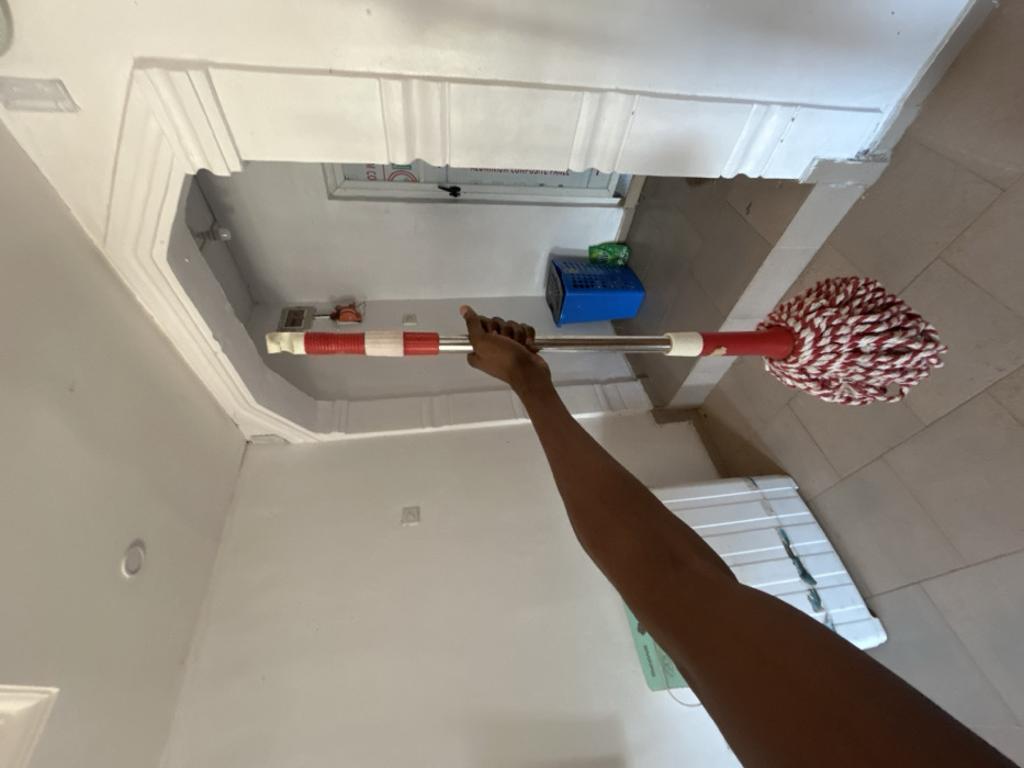 An interior shot in Egbe, Nigeria, depicts a dark-skinned human arm extending from the left, holding a red and white string mop vertically. The mop features a silver metallic handle with red and white banded plastic sections near the top and a cream-colored plastic collar above the red and white string mop head.
The room has white painted walls and a light-colored tiled floor. A recessed ceiling light is visible in the upper left. To the left, a white appliance, possibly a refrigerator or freezer, is partially visible, topped with a light green object.
A decorative white archway with multiple moldings and columns dominates the central-right portion of the frame, leading to an adjacent area with a darker tiled floor. Within this secondary area, a blue plastic basket is visible on the floor, next to a small green object. A white door with partially visible text is located in the background to the right of the archway. An electrical box with red wiring is mounted on the wall to the left of this door. The overall scene is brightly lit.