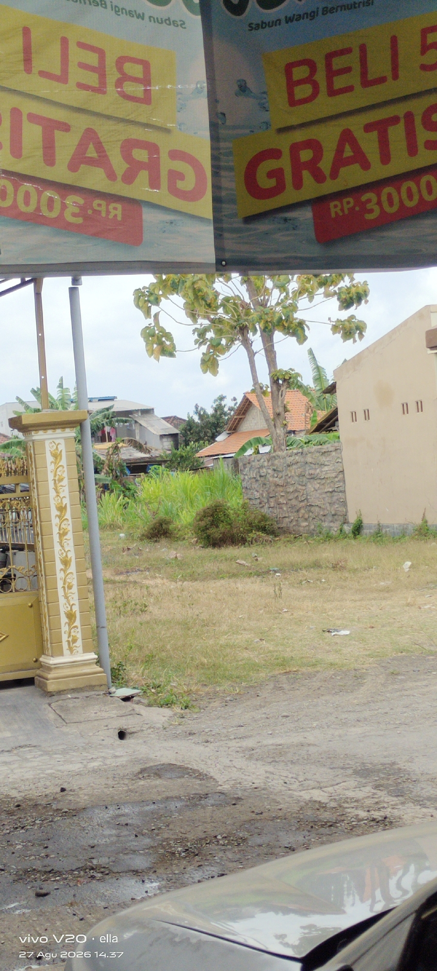 The image displays a street scene in Simo, Indonesia. A banner with Indonesian text, "BELI GRATIS RP. 3000" is visible, indicating a promotional offer. A building with a light-colored facade and dark windows is present. Adjacent to it is a stone wall and lush green vegetation. A large tree with sparse leaves stands in the foreground. A gate with decorative elements and a pillar are also visible on the left side of the image. The ground is unpaved with patches of dry grass and debris. The sky is overcast. The timestamp "vivo V20 · ella 27 Agu 2026 14.37" suggests the photo was taken on August 27, 2026.