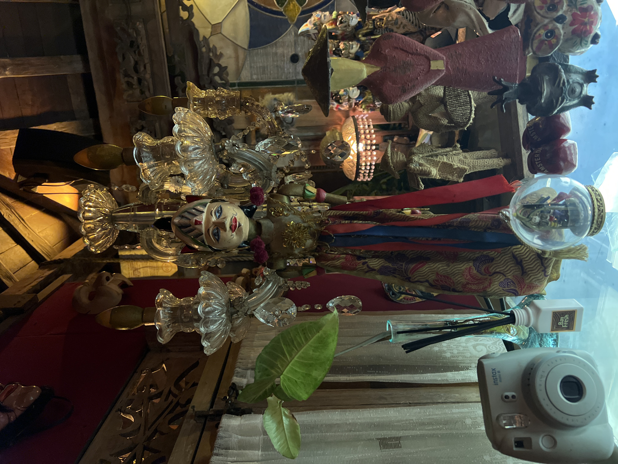 The image shows a cluttered interior, likely in North Kuta, Indonesia. A prominent decorative chandelier with a painted face hangs in the center. Various objects surround the chandelier, including decorative figures, textiles, and a glass snow globe. In the foreground is a green plant and a white Instax Mini camera. The environment appears to be an eclectic mix of decorative items with a warm, dim lighting.