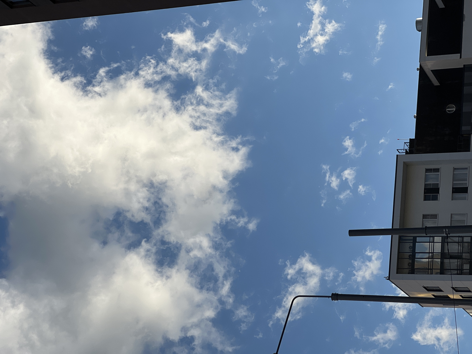 The image depicts a view of the sky and the upper floors of modern buildings, likely in Yekaterinburg, Russia. The sky is a vibrant blue, with scattered white cumulus clouds, some appearing fluffy and others more wispy. A tall, dark grey lamppost with a curved arm is visible on the left side of the frame. To its right, a portion of a white building with large, reflective windows is seen, interspersed with smaller, rectangular windows. Behind this white section, a dark-colored section of another building is present. Two tall, slender, dark grey cylindrical structures, possibly ventilation shafts or decorative elements, stand in front of the buildings. The top edges of several buildings are visible, indicating an urban setting. No people are discernible in the image. The lighting suggests it is daytime, with sunlight illuminating the clouds and buildings.