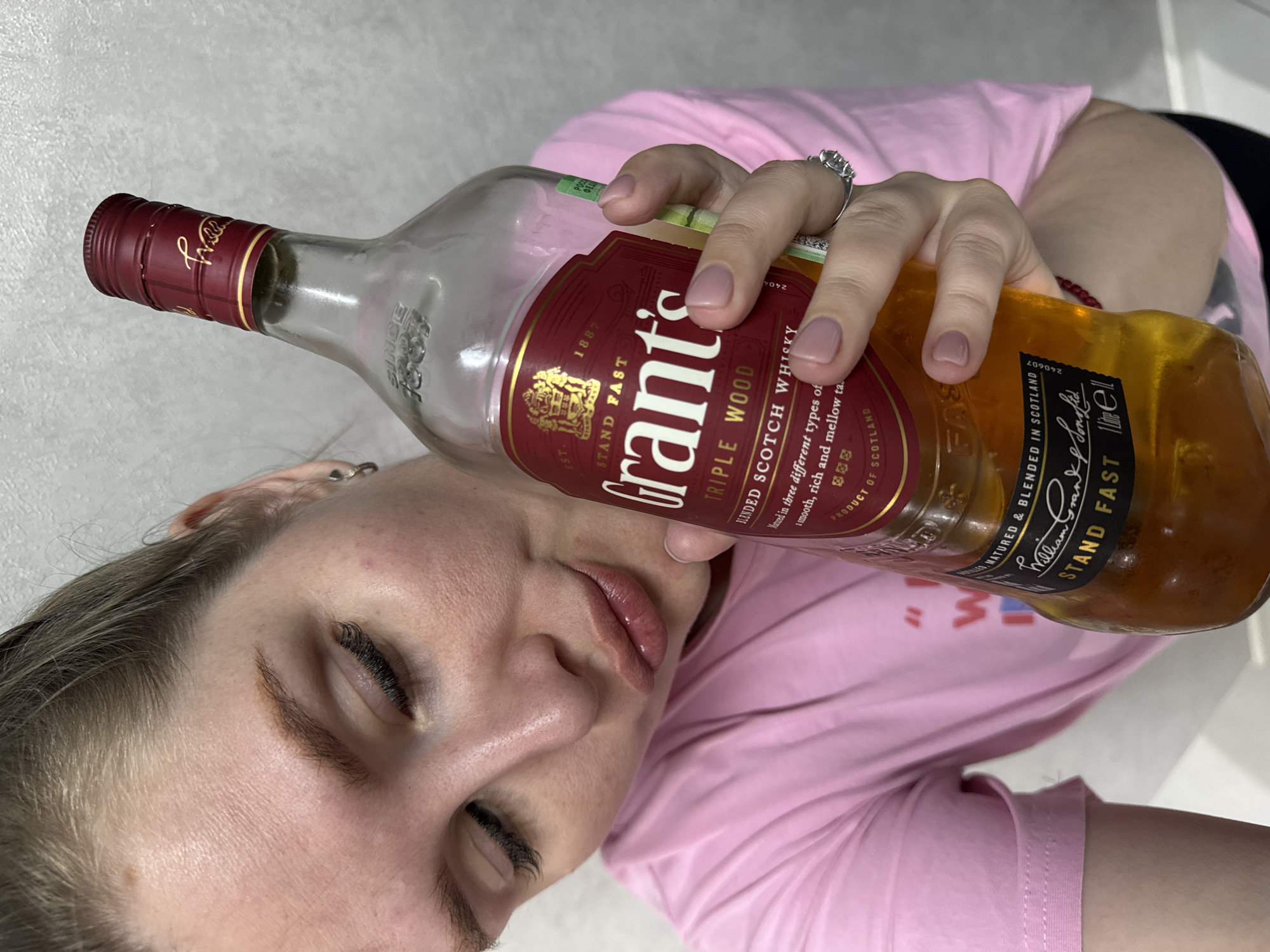 A woman with fair skin and closed eyes is seen holding a bottle of Grant's Triple Wood Blended Scotch Whisky against her cheek. She is wearing a pink t-shirt and a ring on her finger. The label of the bottle is visible, displaying the brand name, type of whisky, and other information. The image's location is listed as Gornyi (Dvurechenskoe S/P), Russia.