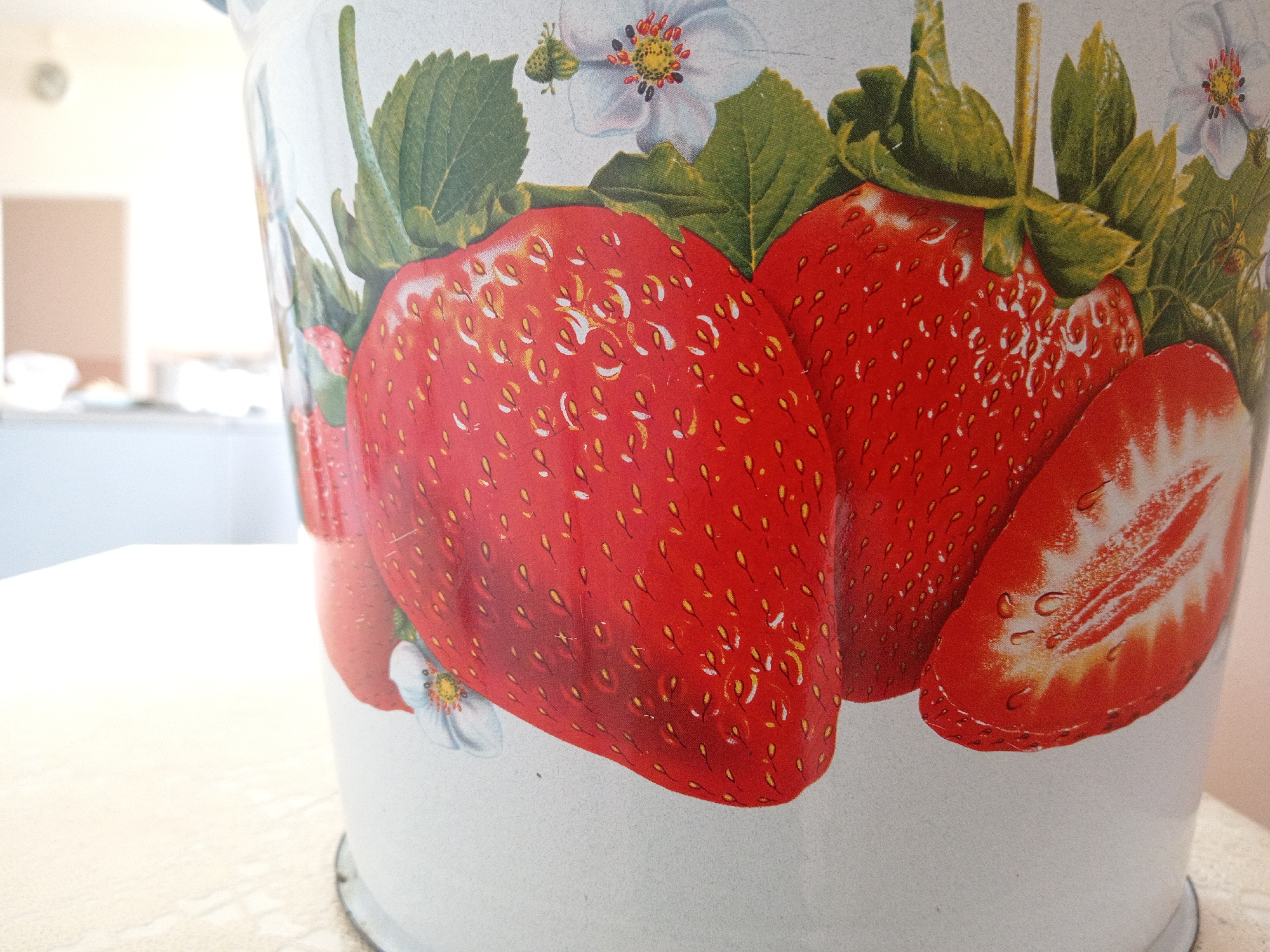 The image shows a close-up of a white metal bucket with a strawberry pattern. The strawberries are depicted in bright red with yellow seeds and green leaves. There are also white flowers with red centers interspersed among the strawberries. The bucket has a slightly curved surface, and the pattern wraps around it. The background is out of focus and appears to be an indoor setting with a light-colored wall and possibly a doorway or window. The lighting is soft and diffused, suggesting it might be daylight. The image does not contain any people, nor does it provide any specific information about its location in Uchkeken, Russia.