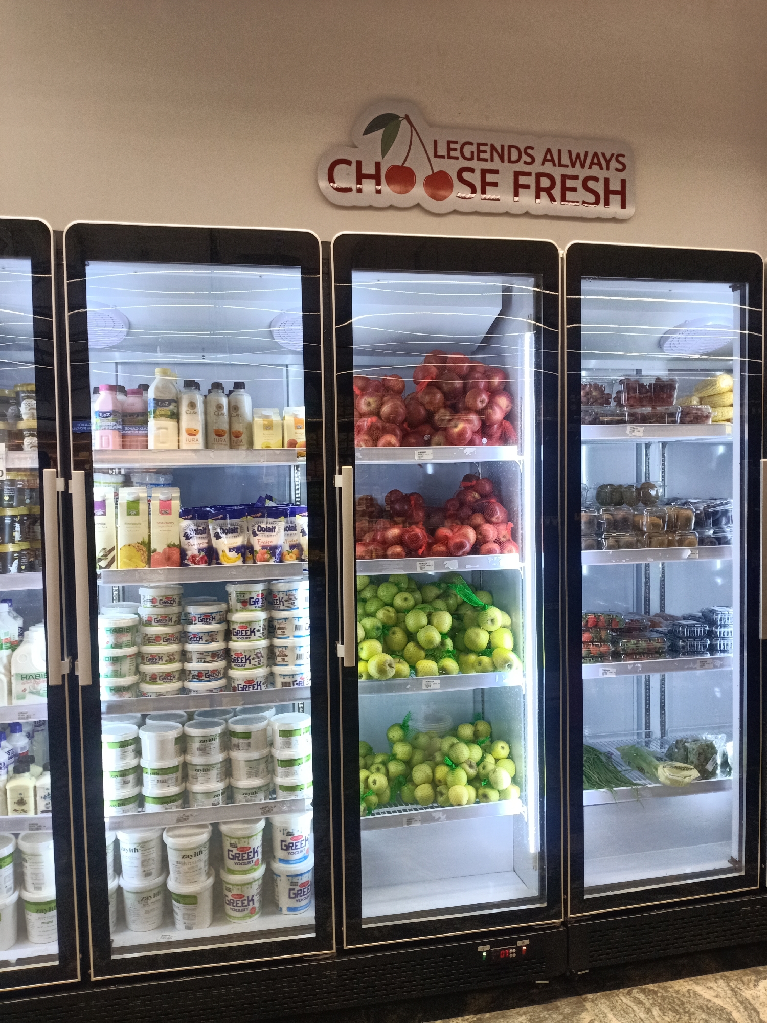 The image shows refrigerated display cases stocked with fresh produce and dairy products, likely in a grocery store or market. The first case on the left contains various dairy items, including yogurts and drinks. The second case is filled with red and green apples, some packaged in bags. The third case contains packaged grapes, strawberries, and leafy green vegetables. Above the cases, a sign reads "LEGENDS ALWAYS CHOOSE FRESH" with cherry images. The environment suggests a retail setting focused on providing fresh food options.