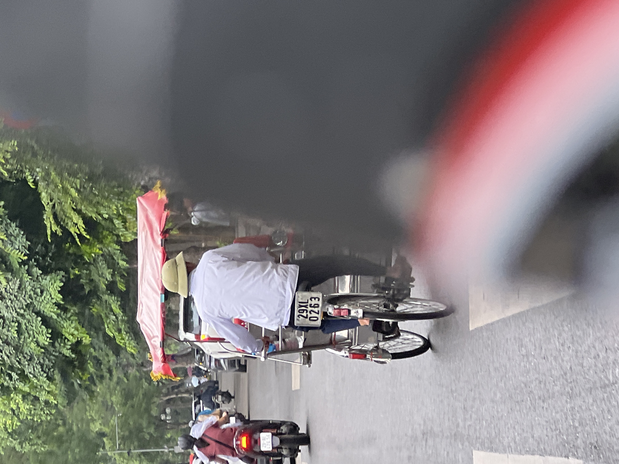 In a street scene in Hanoi, Vietnam, a person wearing a white long-sleeved shirt and a light green bucket hat operates a cyclopousse, a three-wheeled passenger vehicle. The cyclopousse has a red canopy and the license plate reads "29XL 0263". The driver is positioned behind the passenger compartment, which is covered by the canopy. The vehicle is on a paved road, with other vehicles, including motorcycles and cars, visible in the background. Trees line the side of the road. The image is taken from the perspective of a person on a motorcycle, with the front of their vehicle, including a blurred red and white stripe, obscuring part of the right side of the frame. The sky appears overcast.