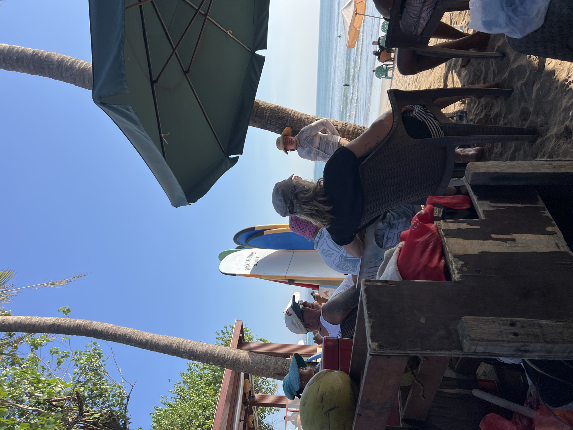 On a sunny day at Kuta Beach, Bali, Indonesia, a group of people are gathered under a large green umbrella near a palm tree. In the foreground, a wooden cart holds a large green coconut and some red fabric. Several individuals are seated in plastic chairs, facing the ocean. Two surfboards, one with the logo "BALIGUNS SURF," are propped up on the sand. Further back, more beachgoers are visible, some sitting and others walking. The ocean is calm with small waves and a few surfers can be seen in the distance. The atmosphere appears relaxed, with people enjoying the beach and the shade.