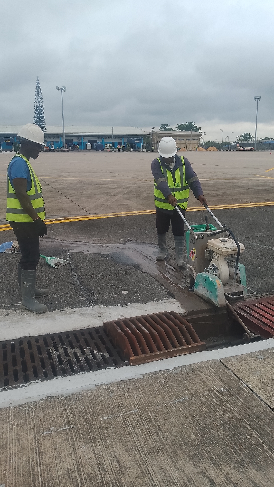 Two male workers in safety gear are repairing an asphalt surface next to a metal drain grate. One worker operates a concrete saw while the other stands nearby with a shovel. They both wear white hard hats, black gloves, and high-visibility vests over their clothing, along with rubber boots. The grate is reddish-brown and borders a concrete surface treated with white paint. Beyond the workers, the area stretches into a paved tarmac with yellow lane markings, leading to a building. The scene appears to be on the grounds of the Warri Airport in Orerokpe, Nigeria, given the airport structures in the background and the presence of the tarmac. The weather appears overcast and dull.
