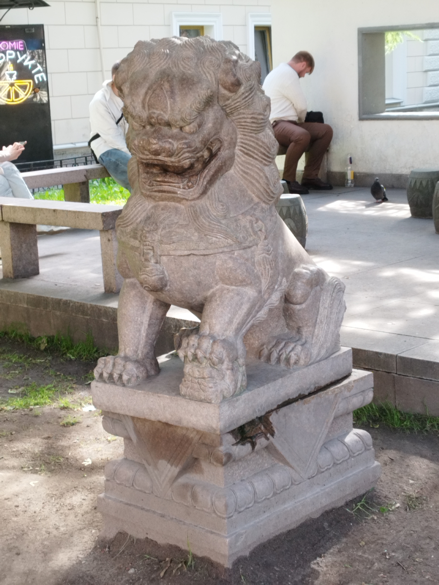 In an outdoor urban setting, likely Saint Petersburg, Russia, a stone statue of a lion dog, characteristic of East Asian designs, stands prominently in the foreground. The statue is weathered and rests on a stone pedestal adorned with decorative carvings. To the left of the statue, a bench with a stone slab seat is visible, with a person partially obscured by the lion dog, seated and looking at their phone. Further left, a black sign with neon lettering in an unfamiliar script and a graphic of a yellow half-circle with radiating lines is mounted on a white building. In the middle ground, a man with reddish-brown hair, wearing a white long-sleeved shirt and brown pants, is seated on a bench, looking down. A dark bag rests beside him. To the right of the seated man, a single pigeon stands on a paved surface, with another stone object partially visible behind it. The ground is a mix of pavement and soil with sparse green vegetation. The background shows a white building with windows and architectural details. Sunlight casts shadows across the scene, indicating it is daytime.