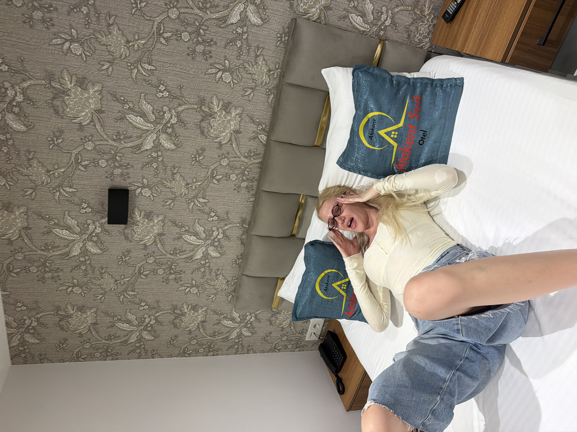 A high-angle shot reveals a woman lying on a bed in a room likely located in Küçükçekmece, Turkey. The room features a grey patterned wallpaper. The woman, wearing glasses, a light-colored top, and denim shorts, has her hands raised near her face. The bed is made with white linens and two blue decorative pillows with the inscription "Atakent Suit Otel". A telephone and a wooden nightstand are adjacent to the bed. A remote control rests on the nightstand.