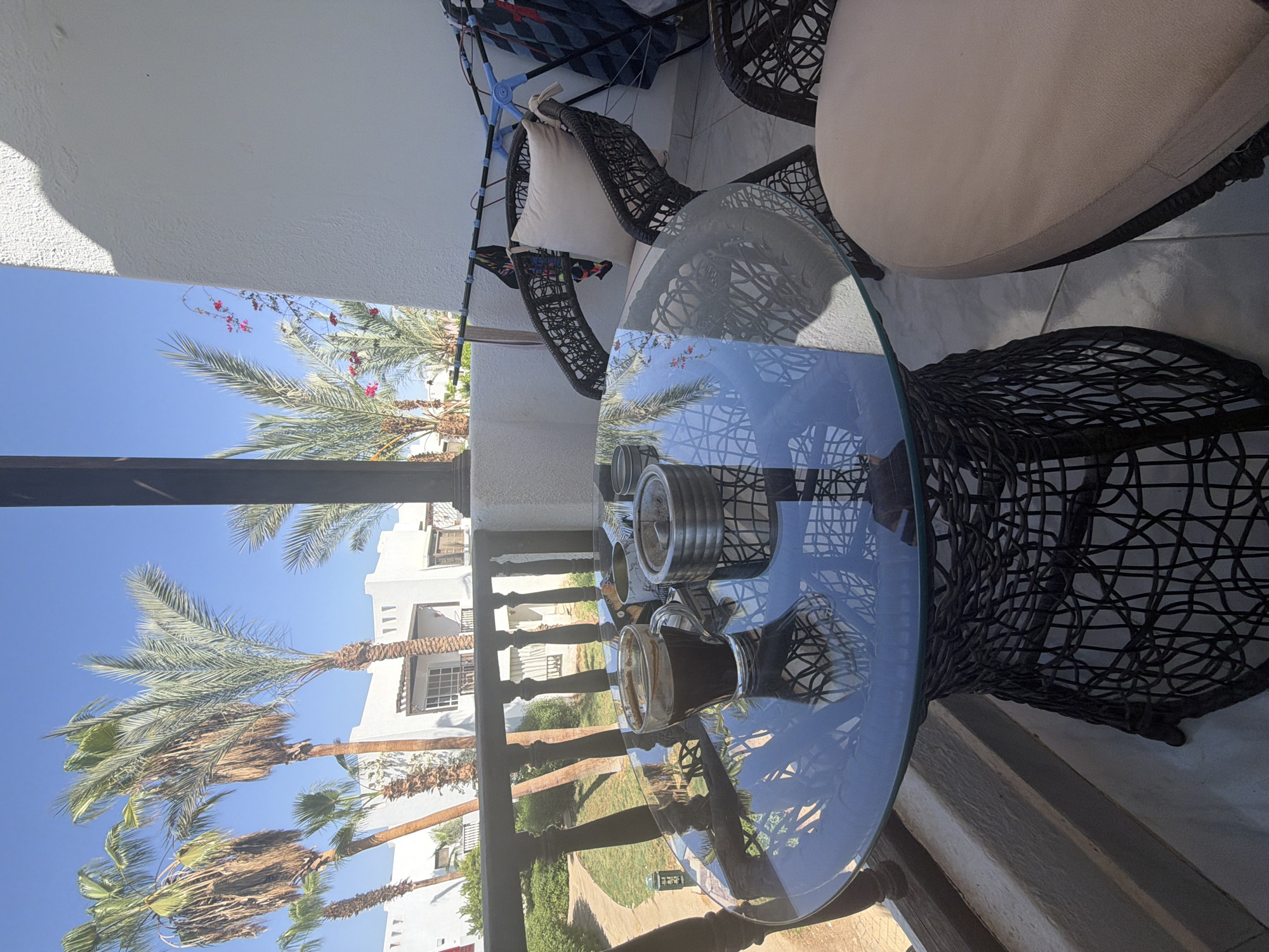 A glass-top table with a woven base is set on a balcony in Sharm El Sheikh, Egypt. On the table sits a clear glass mug filled with dark liquid, likely coffee, and a metal ashtray. Two woven chairs are positioned around the table, one of which has a light-colored cushion. In the background, palm trees sway under a clear blue sky, with white buildings and a pathway visible. The balcony railing is made of dark wood. A clothes drying rack with items of clothing is visible on the right side of the balcony. The overall scene depicts a relaxed outdoor setting.