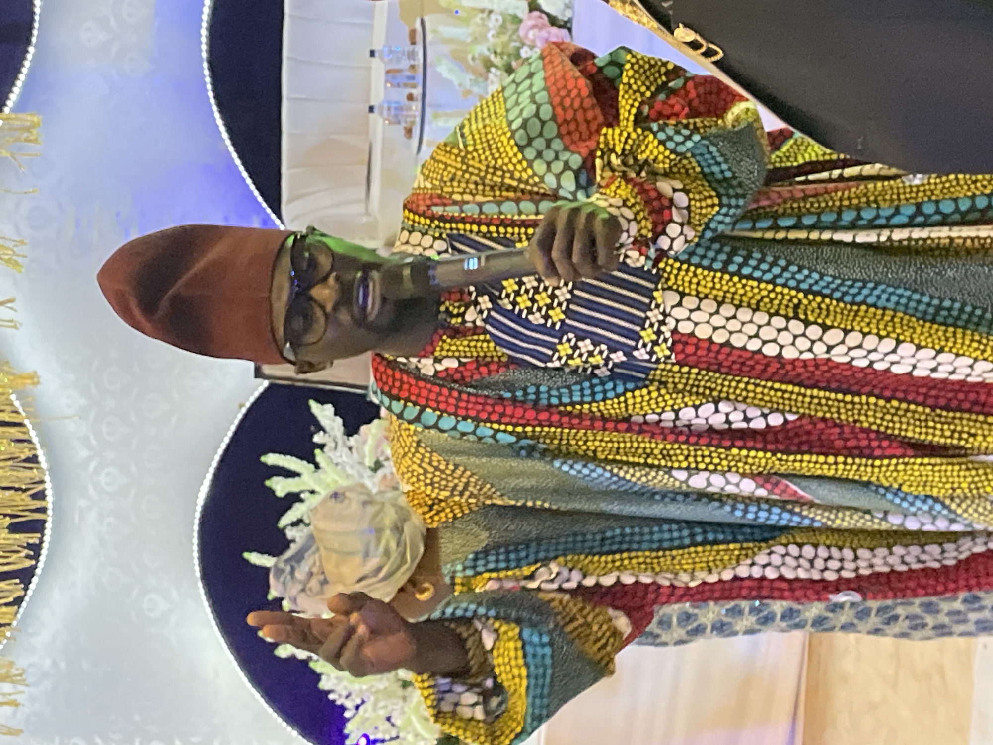 A man is speaking into a microphone, wearing glasses, a brown hat, and a colorful patterned robe. He is gesturing with his left hand. The background includes decorative lighting, fabric, and floral arrangements. The specific location and city of Jos, Nigeria, cannot be definitively verified from the visual information provided.
