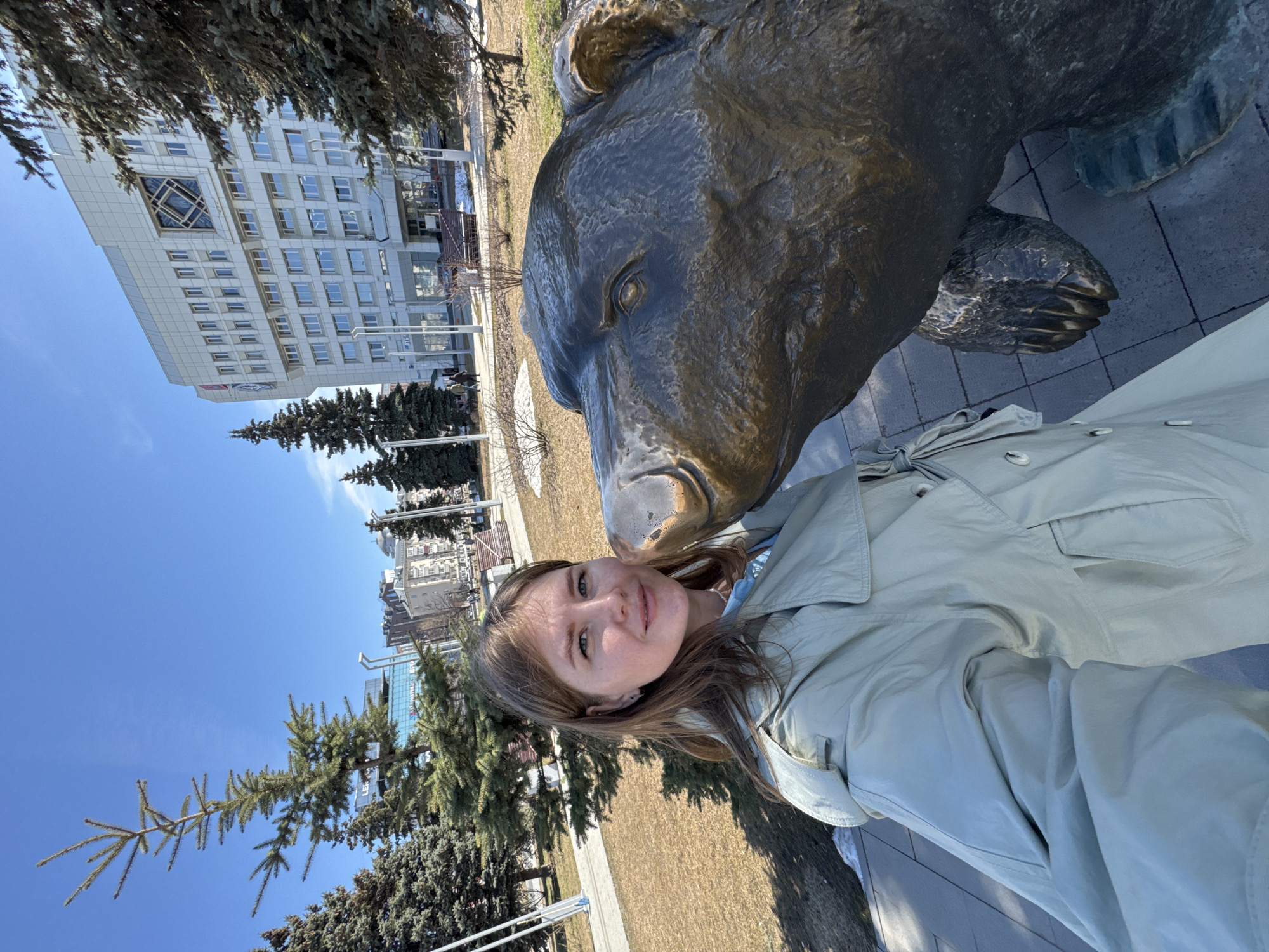 A woman stands next to a large bronze bear statue in Perm, Russia, as she takes a selfie. The bear's nose appears to touch her cheek. In the background are city buildings and evergreen trees under a clear blue sky.