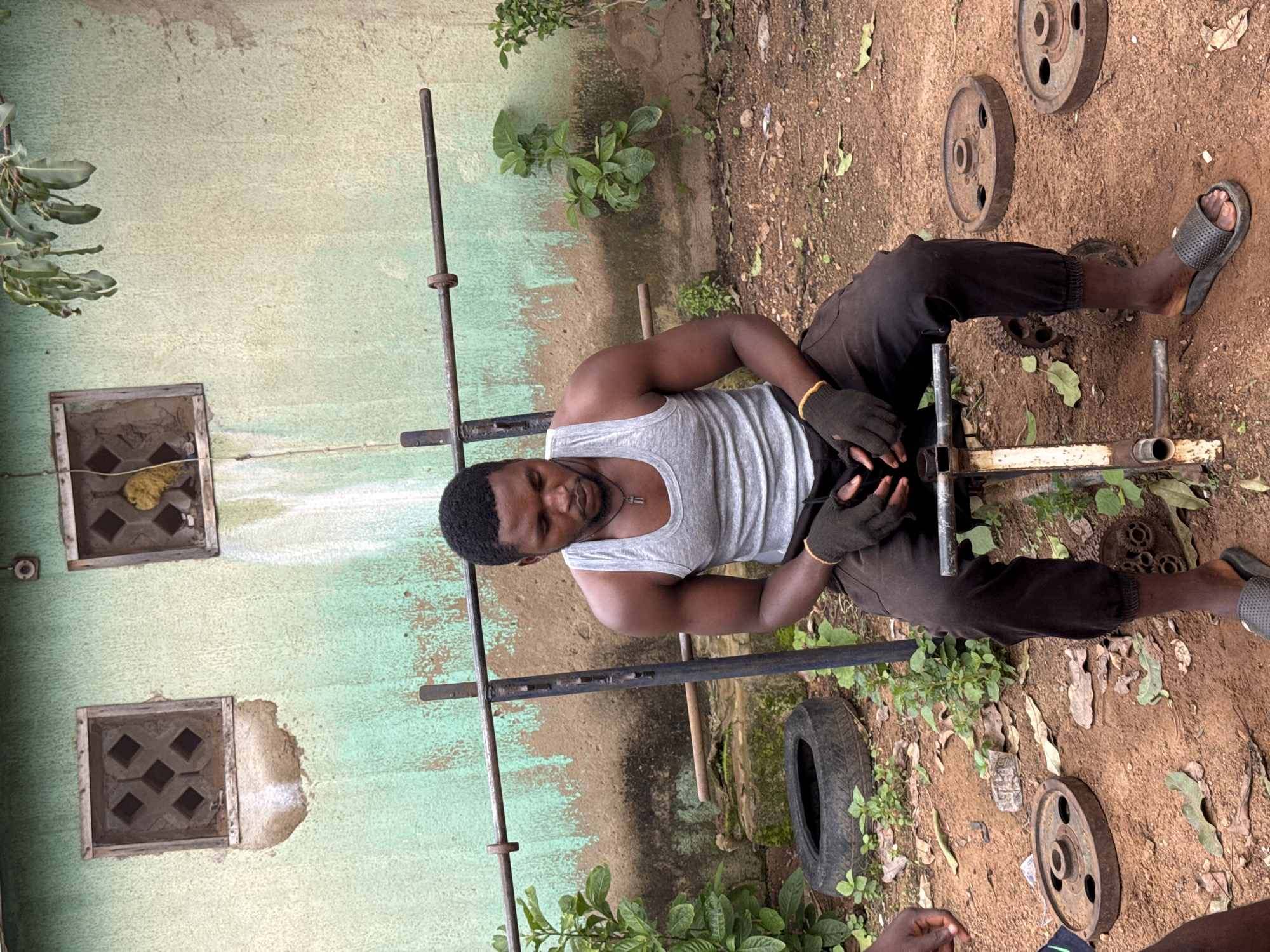 In Akwanga, Nigeria, a Black man wearing a grey tank top and dark pants sits on a weight bench. He is wearing black fingerless gloves and has a silver pendant on a cord around his neck. To his left, a car tire and some green foliage are visible. To his right and in the foreground, several weight plates are scattered on the dirt ground. Behind the man, a barbell rests on a weight rack against a weathered, light green wall with two square window openings. The environment appears to be an outdoor setting.