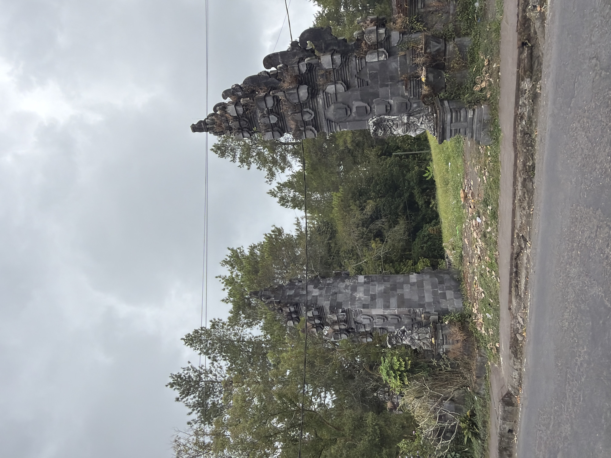 The image depicts a cloudy day in Kintamani, Indonesia, featuring two ornate stone gates. These gates are constructed from dark, carved stone blocks and are adorned with intricate patterns and small statues. One gate is positioned on the left side of the frame, and the other on the right. Between the gates, there is a grassy area bordered by trees. A paved road runs in the foreground, separating the viewer from the gates and the natural landscape. Power lines are visible stretching across the upper portion of the image, against the overcast sky. The overall scene suggests a roadside location with traditional architectural elements set against a backdrop of dense vegetation.