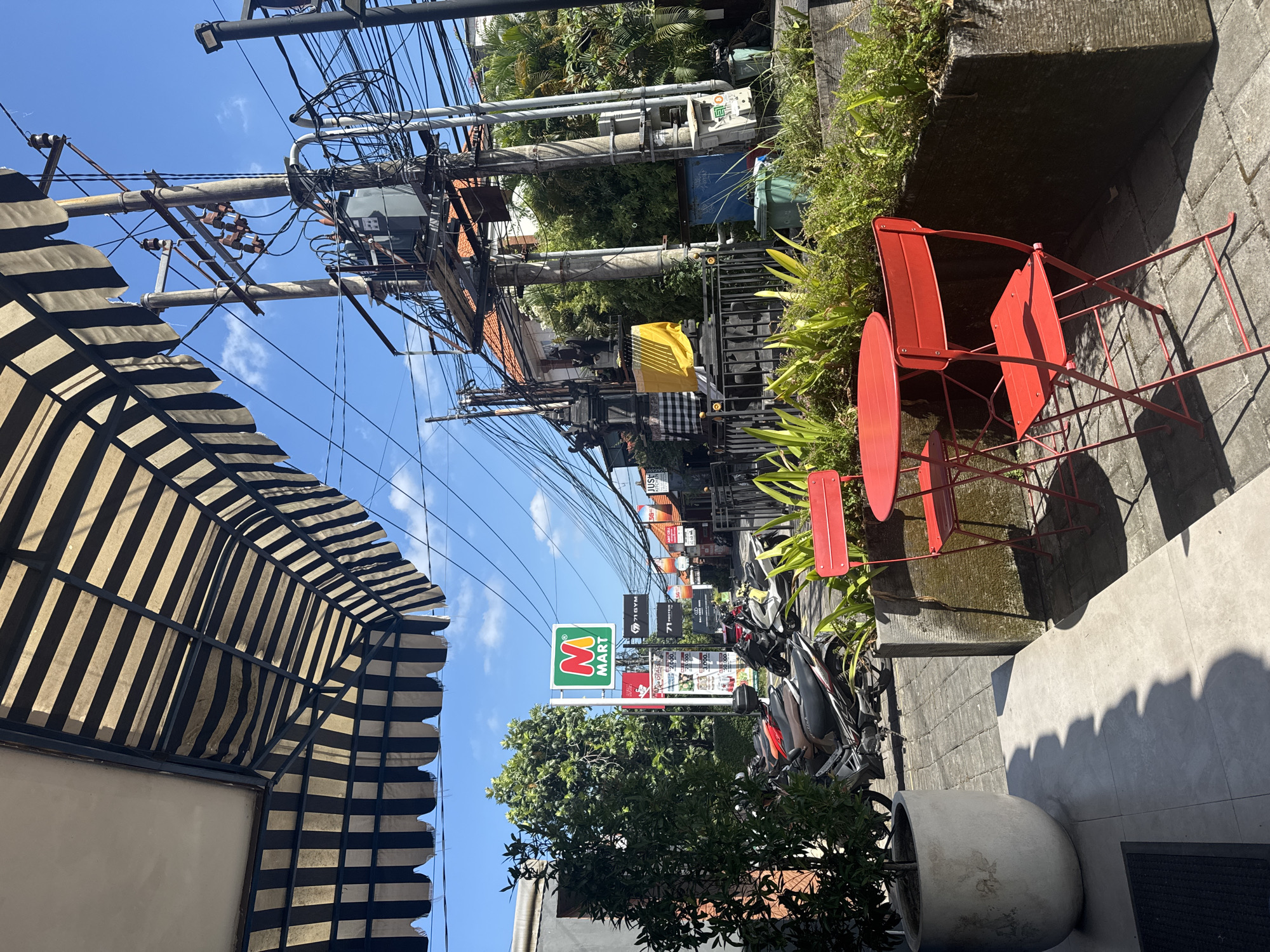 The image depicts a vibrant street scene in North Kuta, Indonesia, under clear, bright sunlight, likely late morning or early afternoon, based on the strong shadows. The perspective is from an outdoor area, possibly a cafe or restaurant, looking down a bustling street.
In the foreground, a prominent black and white striped awning frames the upper left. To the right, a set of two bright red metal chairs and a small matching table are arranged on a light grey concrete patio, suggesting an outdoor seating area. A large cylindrical potted plant sits nearby, and an adjacent raised planter is filled with lush green foliage.
The middle ground showcases typical street infrastructure: numerous utility poles are heavily laden with a dense network of tangled electrical and communication cables, including visible transformers. A large "M Mart" convenience store sign, featuring a green "M" and red "Mart" on a white background, stands prominently on the left side of the street. Further down the street, various commercial signs are visible, with "71 GYM" and "JUST KITCHEN" being discernable. Several motorbikes are parked along the roadside. Cultural elements include a yellow flag and a black-and-white checkered textile, resembling a Balinese poleng cloth, hanging from structures.
The sky is a brilliant blue with a few scattered white clouds, indicating clear and sunny weather. There are no people visible, but the setting suggests an active commercial area.