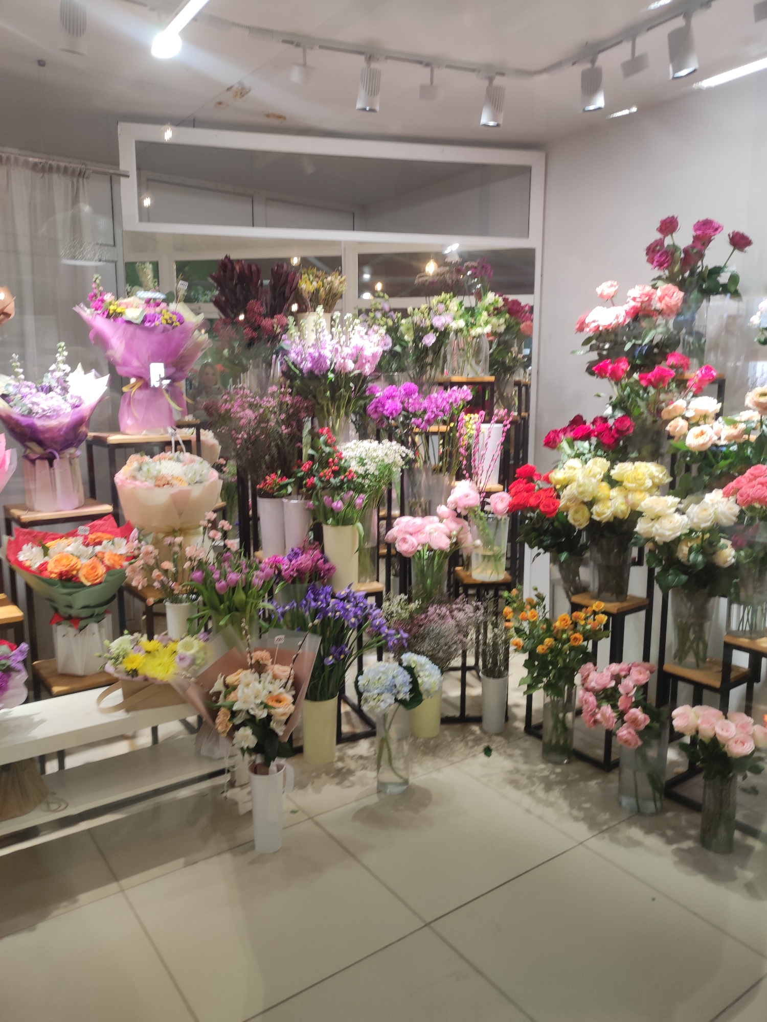 The image shows the interior of a flower shop in Votkinsk, Russia. Various bouquets and floral arrangements fill the space. The flowers include roses, hydrangeas, and other varieties in various colors, arranged in vases and wrapped bouquets. They are displayed on shelves and tables. The flooring is tiled. The lighting is bright and overhead. A large window or storefront can be seen in the background, showing a glimpse of the outside.