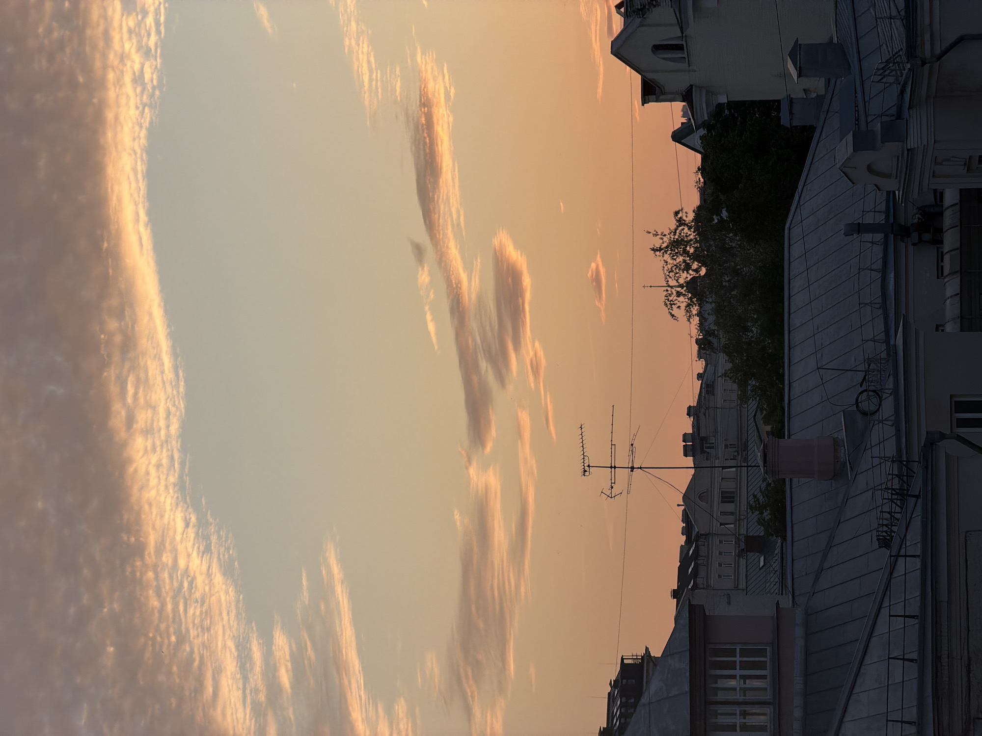 The image depicts a view of a city skyline at sunset, likely in Moscow, Russia, given the architectural style and the presence of a rooftop antenna typical of older European buildings. The sky is a soft gradient of orange and peach, with scattered clouds tinged with pink and gold. Below the sky, the dark silhouettes of buildings and rooftops dominate the lower half of the frame. Rooftops made of metal and tiles are visible, with varying textures and colors, mostly in shades of gray and dark blue. Several chimneys and vents punctuate the rooftops. A prominent television antenna stands in the mid-ground, its metallic structure silhouetted against the sky. Trees, dark and dense, are visible in patches between the buildings. The architecture suggests a dense urban environment with multi-story buildings featuring dormer windows and pitched roofs. There are no people or visible signs of activity. The overall atmosphere is calm and serene, characteristic of a quiet evening in a city.