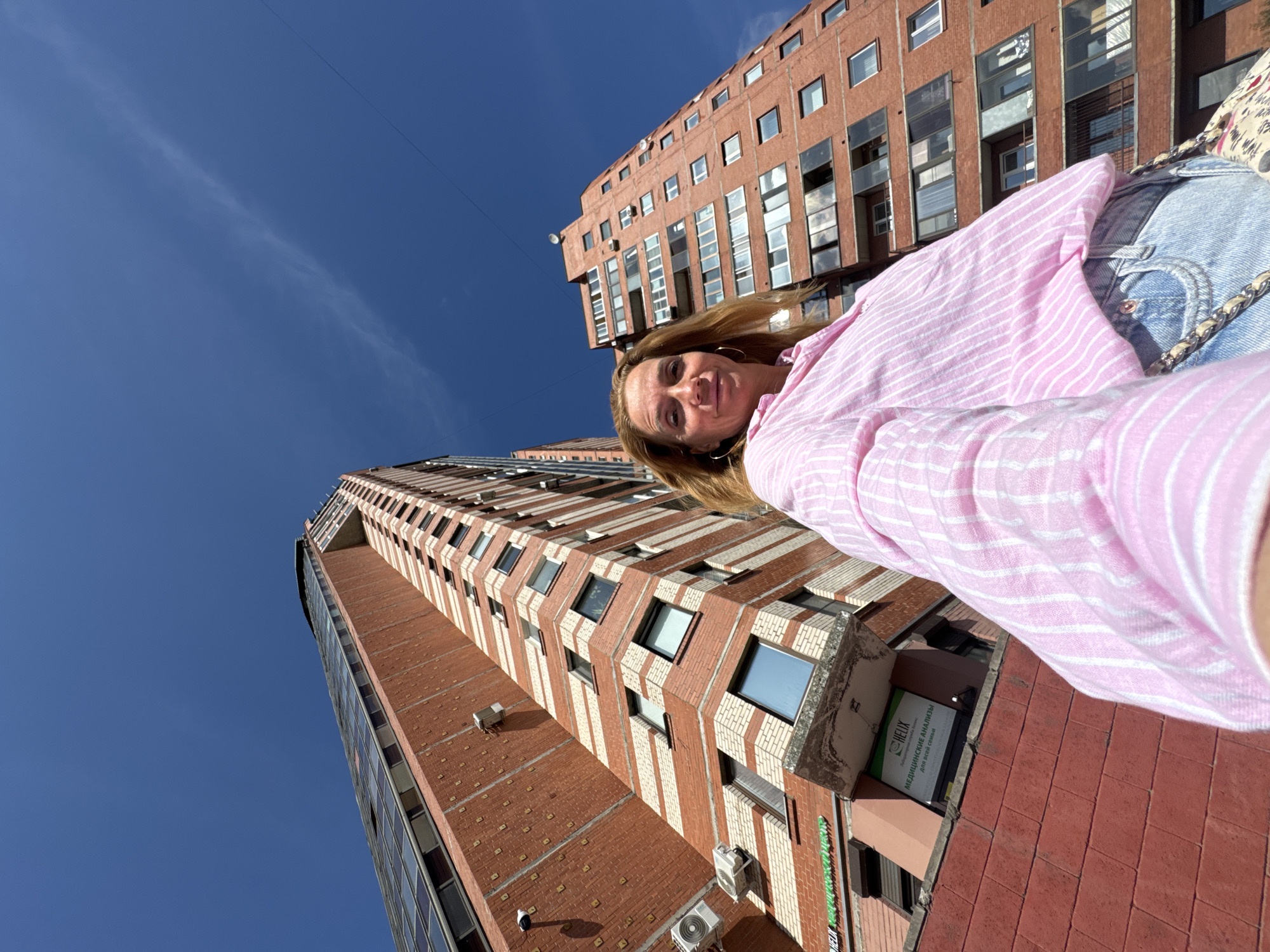 A woman is taking a selfie in front of two brick apartment buildings in Saint Petersburg, Russia. The woman is wearing a pink and white striped shirt and blue jeans. She has blonde hair and is wearing large hoop earrings. The buildings are tall and have many windows. The sky is blue and clear. One of the buildings has a sign that says "HELIX" in green lettering. The overall scene is bright and sunny.