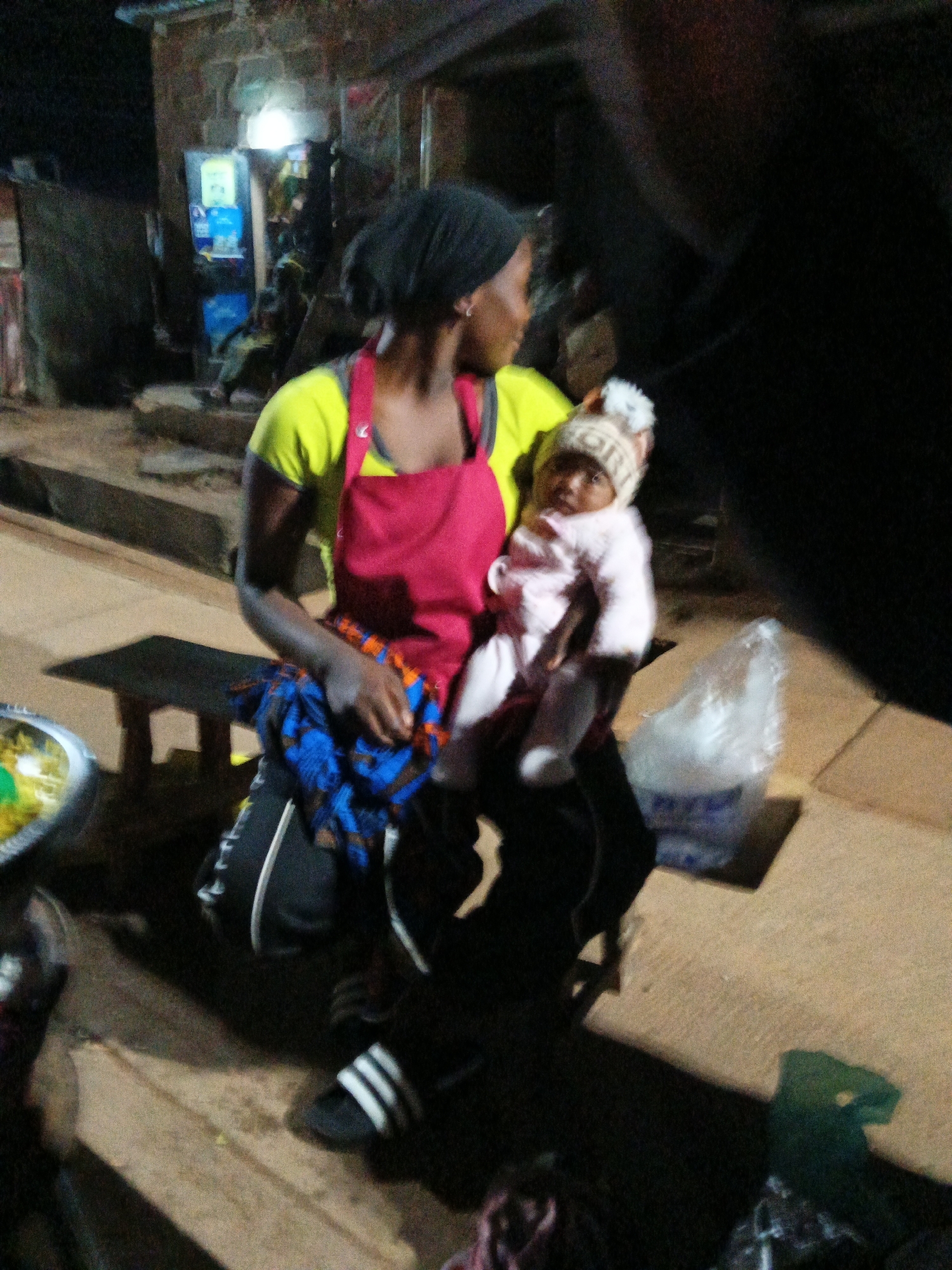 A woman is holding a baby in Bukuru, Nigeria. The woman is wearing a yellow shirt and a red apron. She has a blue and orange fabric draped over her lap and is wearing black pants with white stripes and black and white slide sandals. The baby is wearing a light pink outfit and a knitted hat with a pompom. The scene appears to be outdoors at night, possibly in a market area, with buildings and lighting visible in the background. A plastic bag and a metal bowl are also present in the foreground.